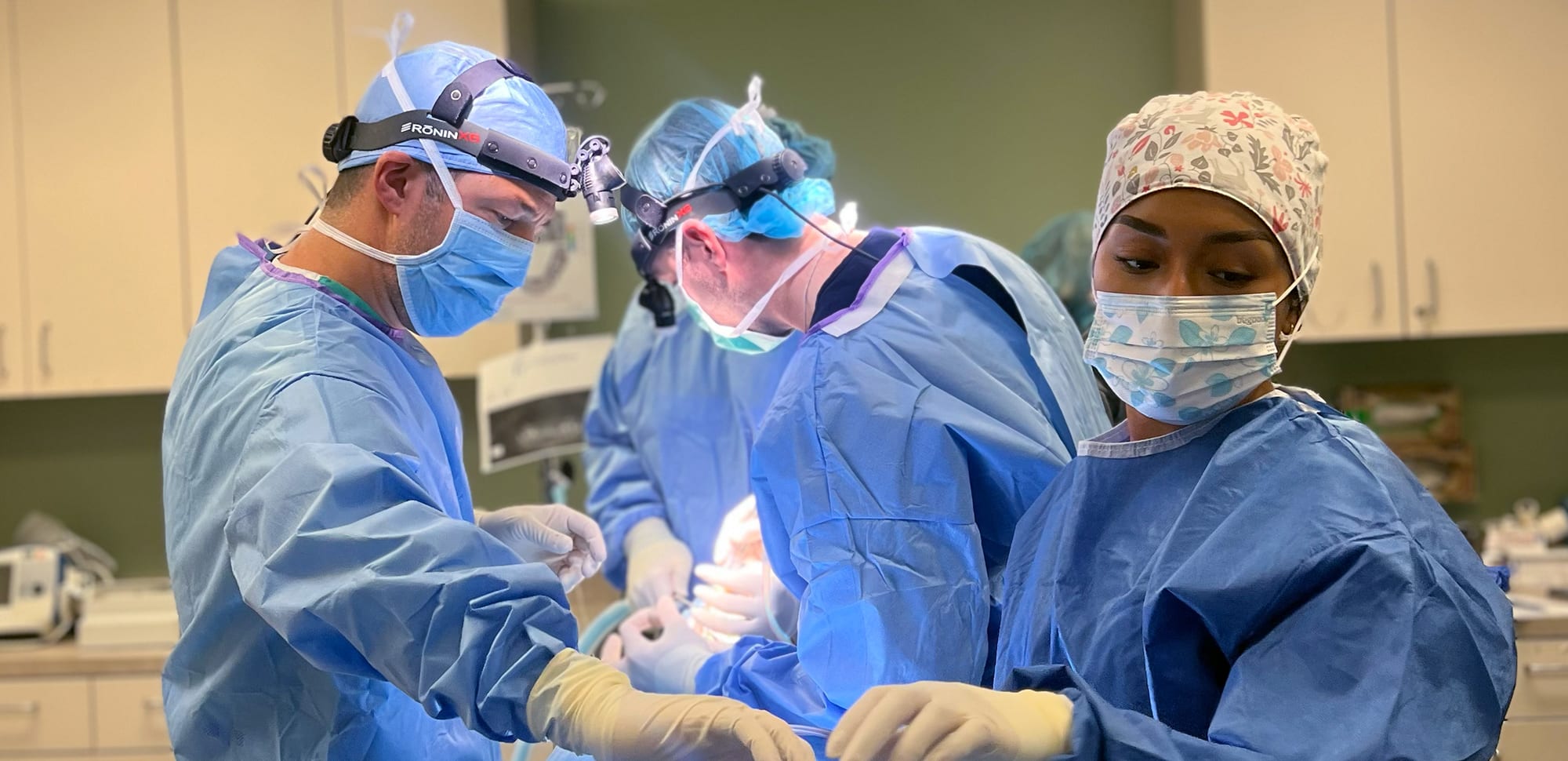 Three medical professionals in an operating room, wearing blue surgical attire and masks, focused on a procedure under bright lights.