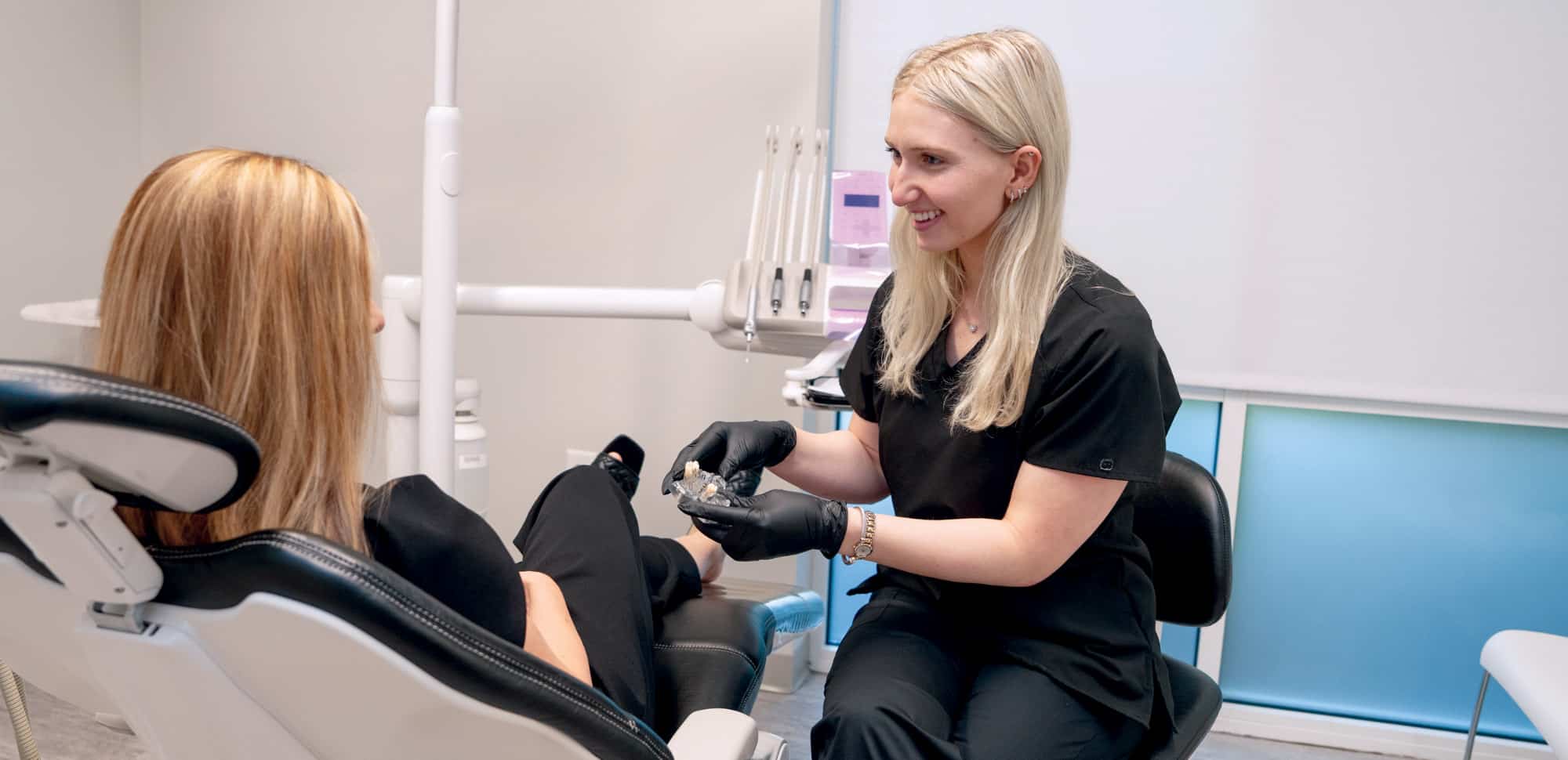 A dental professional shows a clear aligner to a seated person in an examination room with medical equipment around them.