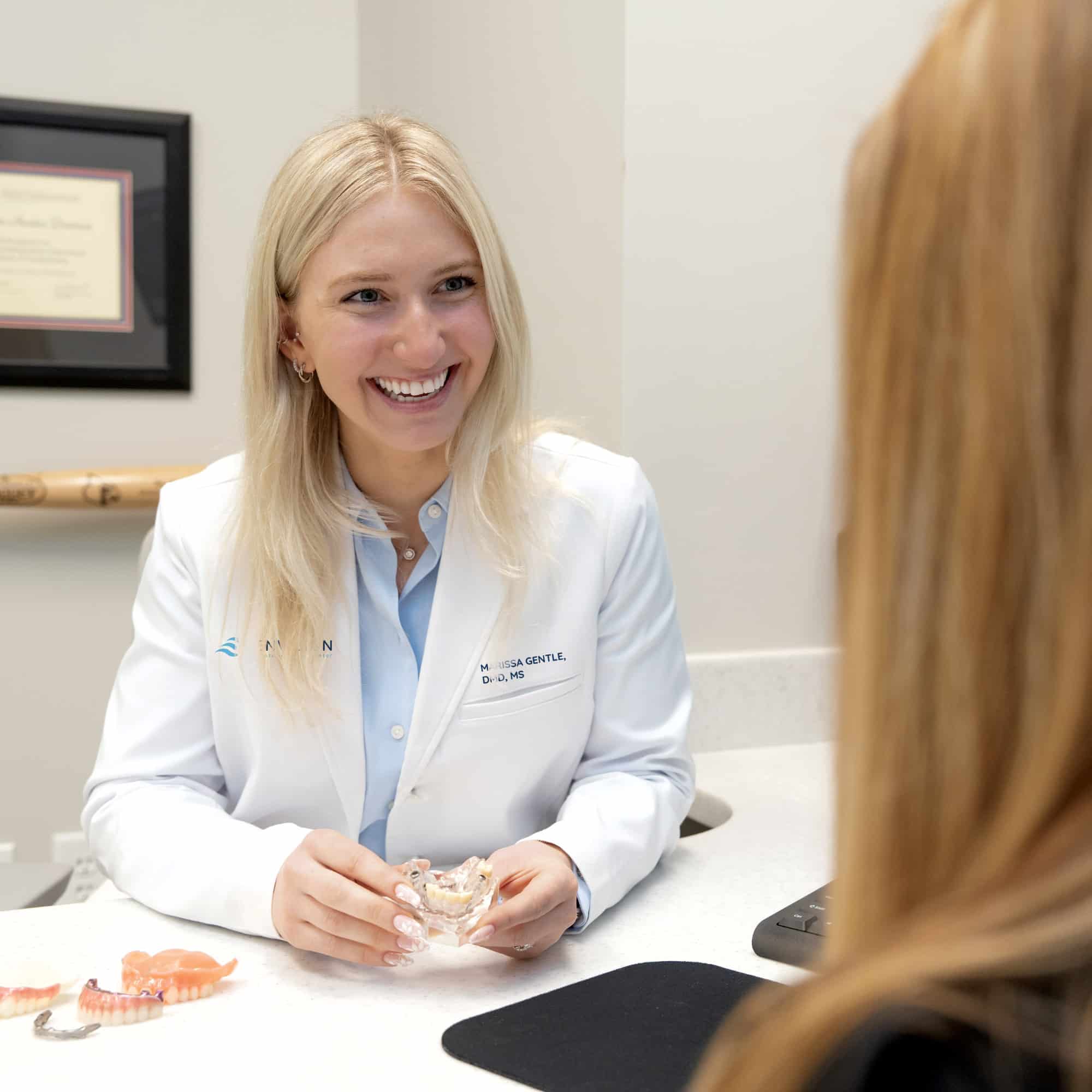 A person in a lab coat smiles while discussing dental models with another person at a desk in an office setting.