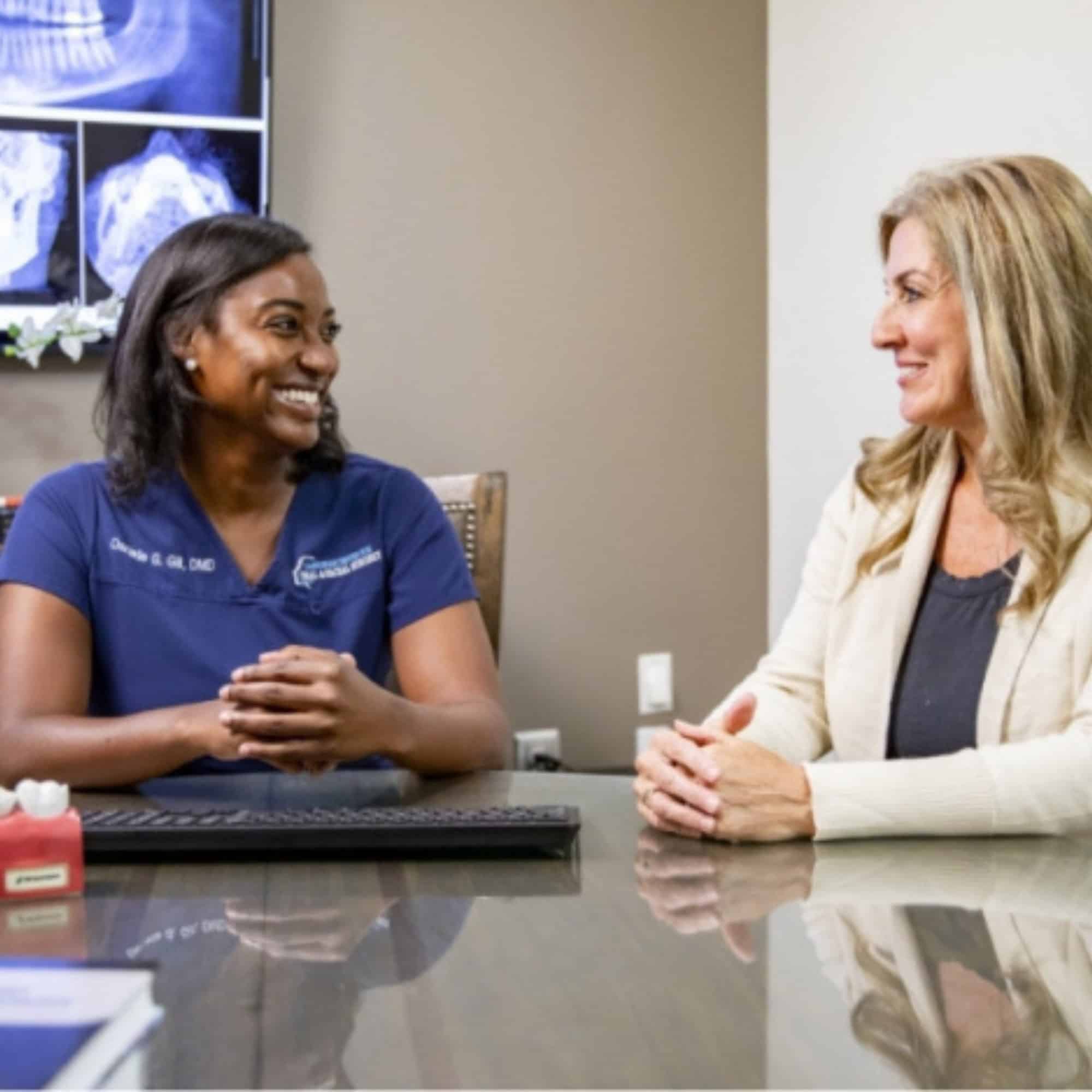 Two people are sitting at a desk, smiling and talking. Dental X-rays are displayed on the screen in the background.