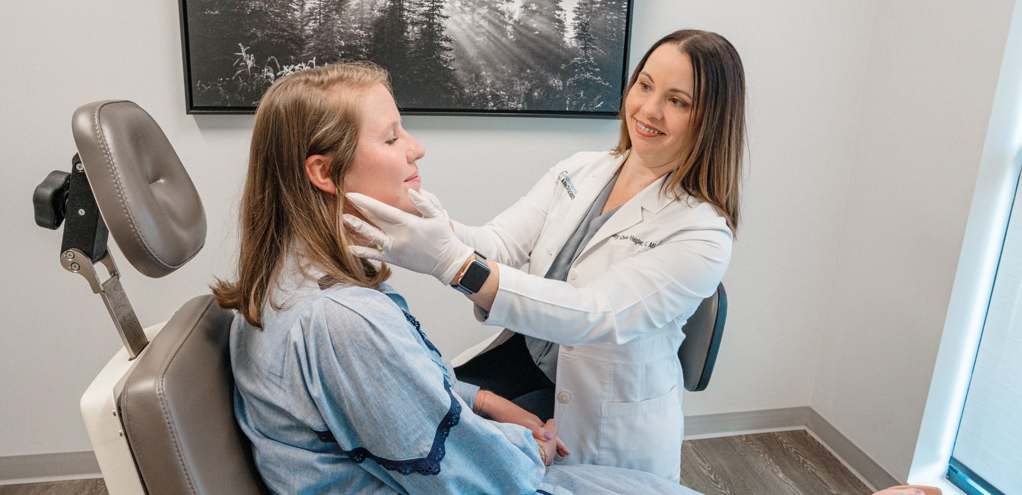 Two people are in a medical office; one examines the other's face. A framed landscape photo is on the wall.