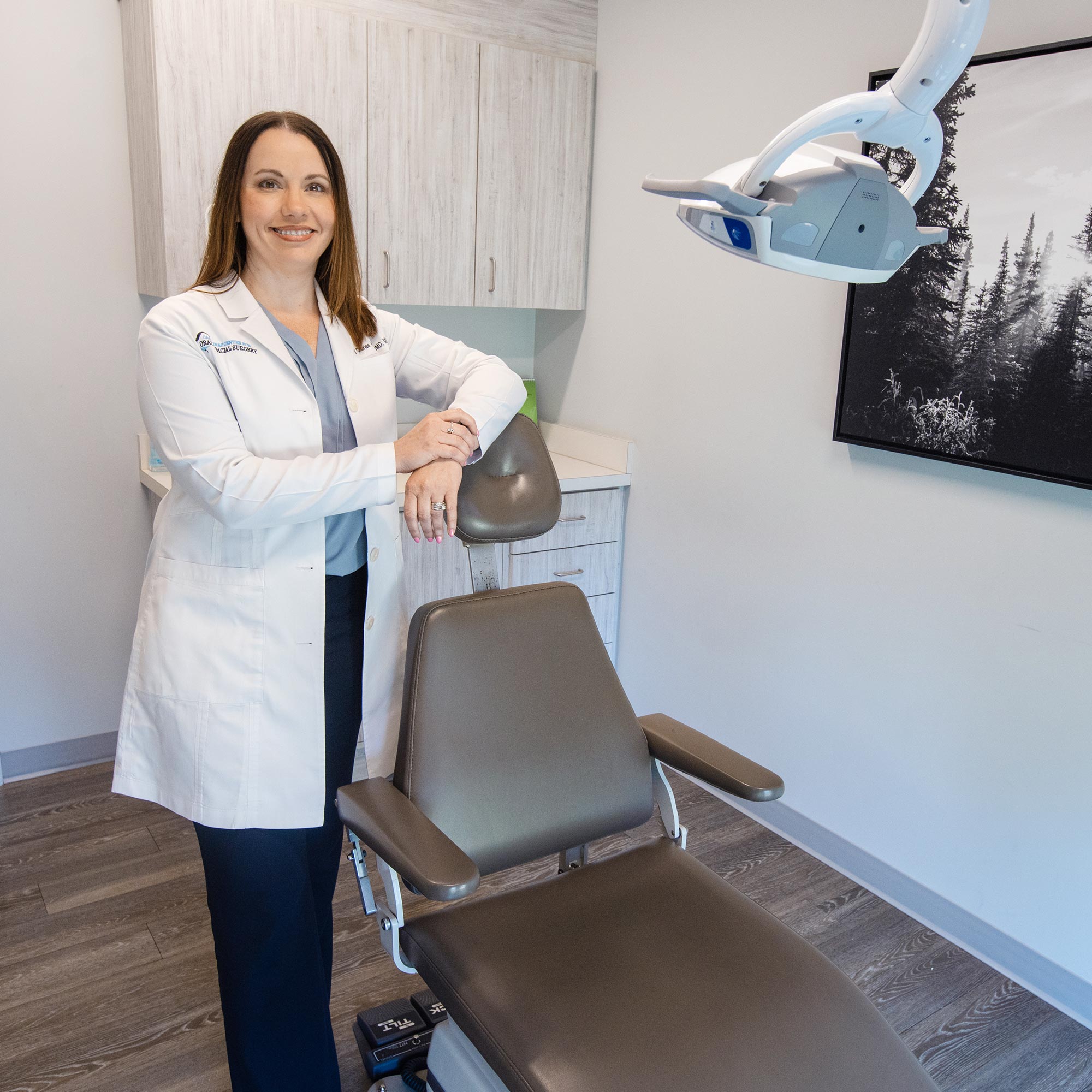 A person in a white coat stands in a modern dental office next to a dental chair, with a nature photo on the wall.