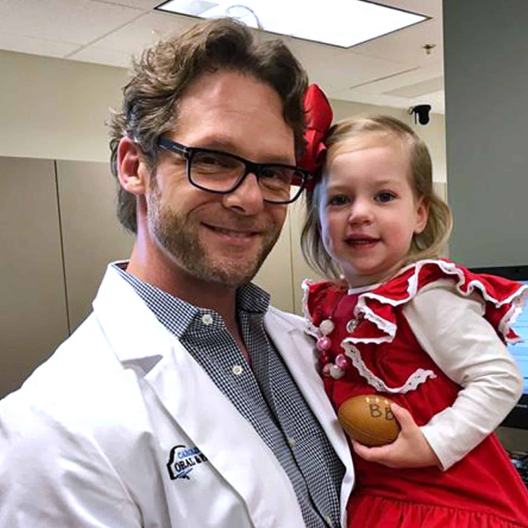 A person in a white coat holds a young child in a red dress indoors, who is holding a small football.
