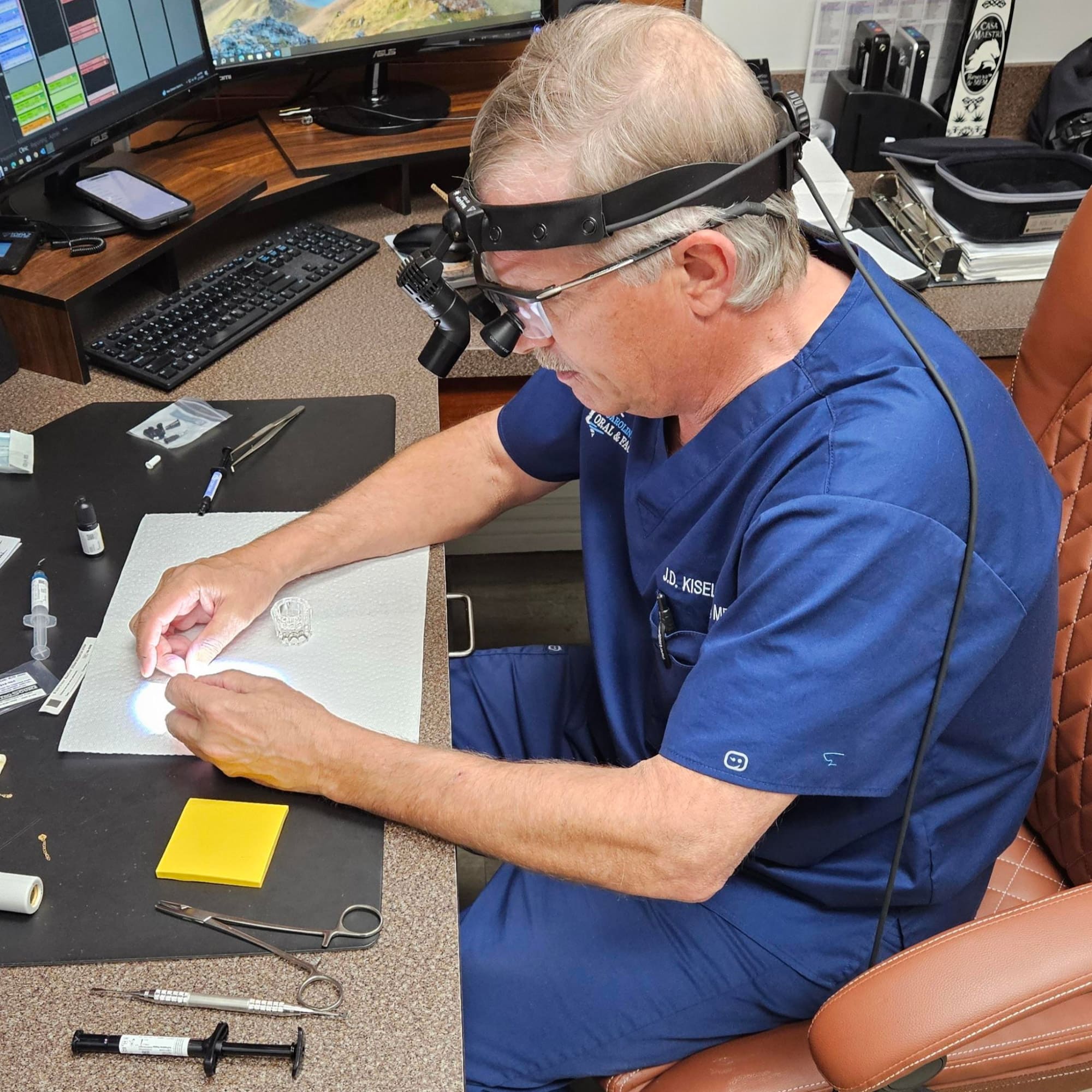 A person in blue scrubs works meticulously with medical tools, wearing magnifying glasses, at a desk with computer and supplies.