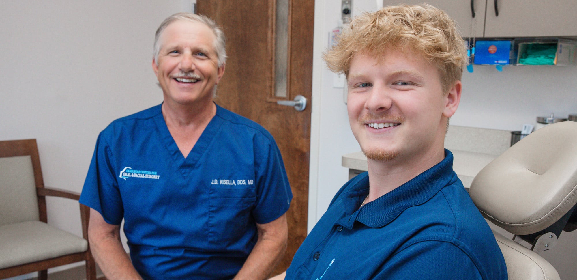 Two persons in blue attire smiling in a clinic setting, seated near medical equipment and a door in the background.