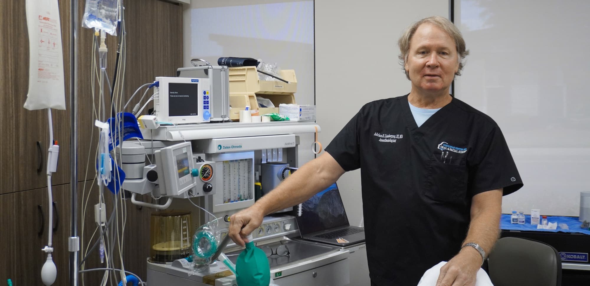 A person in a medical office stands next to anesthesia equipment, wearing scrubs, preparing surgical instruments and supplies.