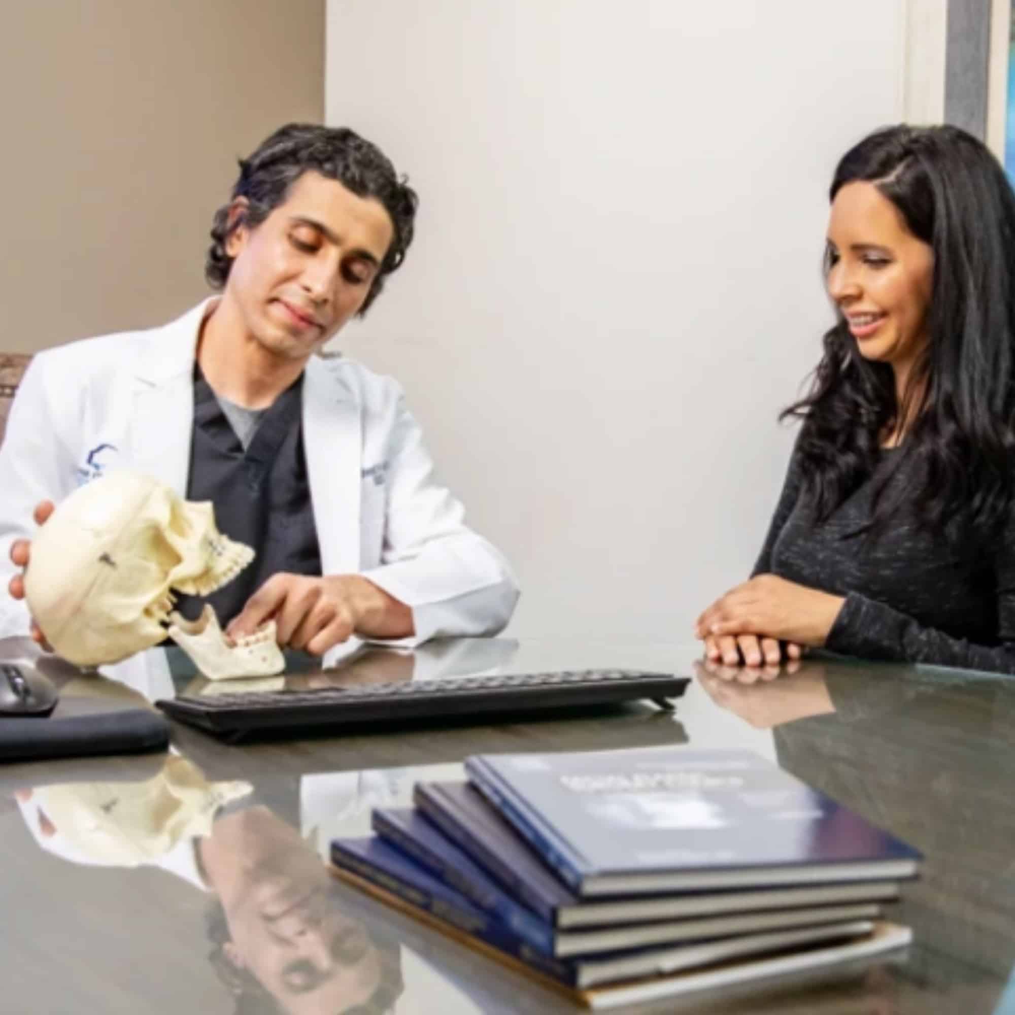 Two people are sitting at a desk with a skull model, having a discussion in a professional office setting. Books are stacked nearby.