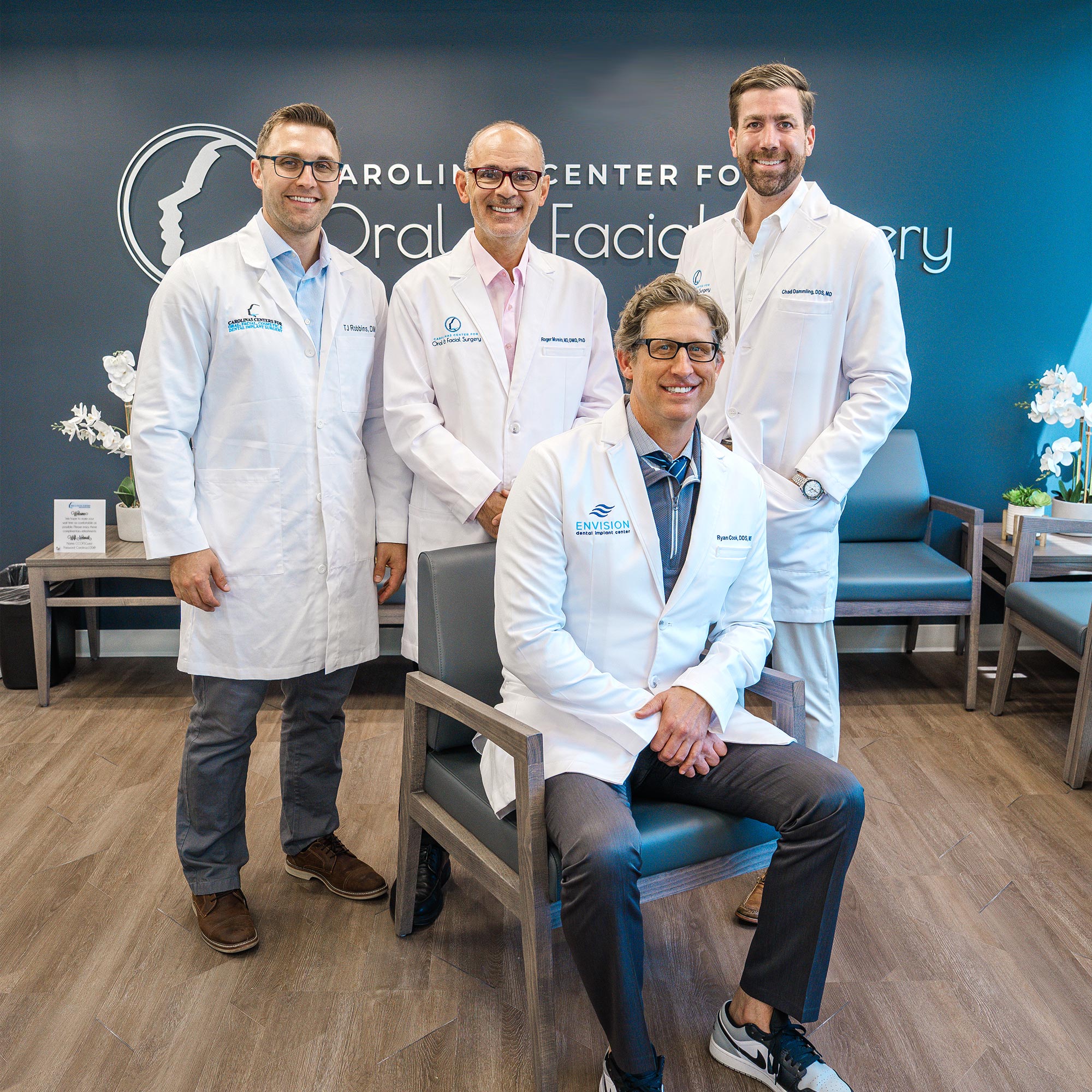 Four people in white coats, smiling, inside an office with a sign reading "Oral Facial Surgery." Modern, professional setting with flowers.