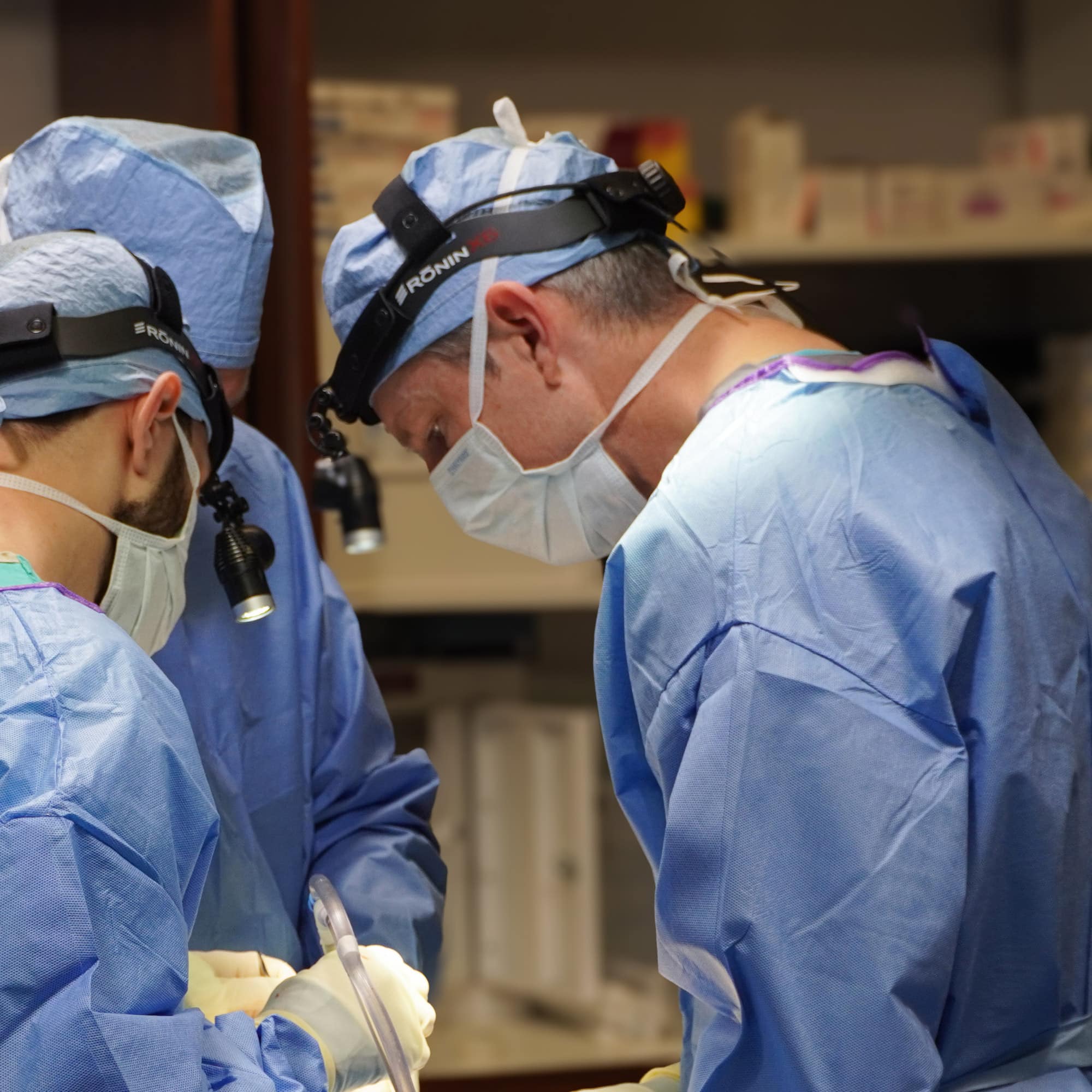 Three people wearing surgical attire and headlamps focus intently on a medical procedure in a hospital operating room setting.