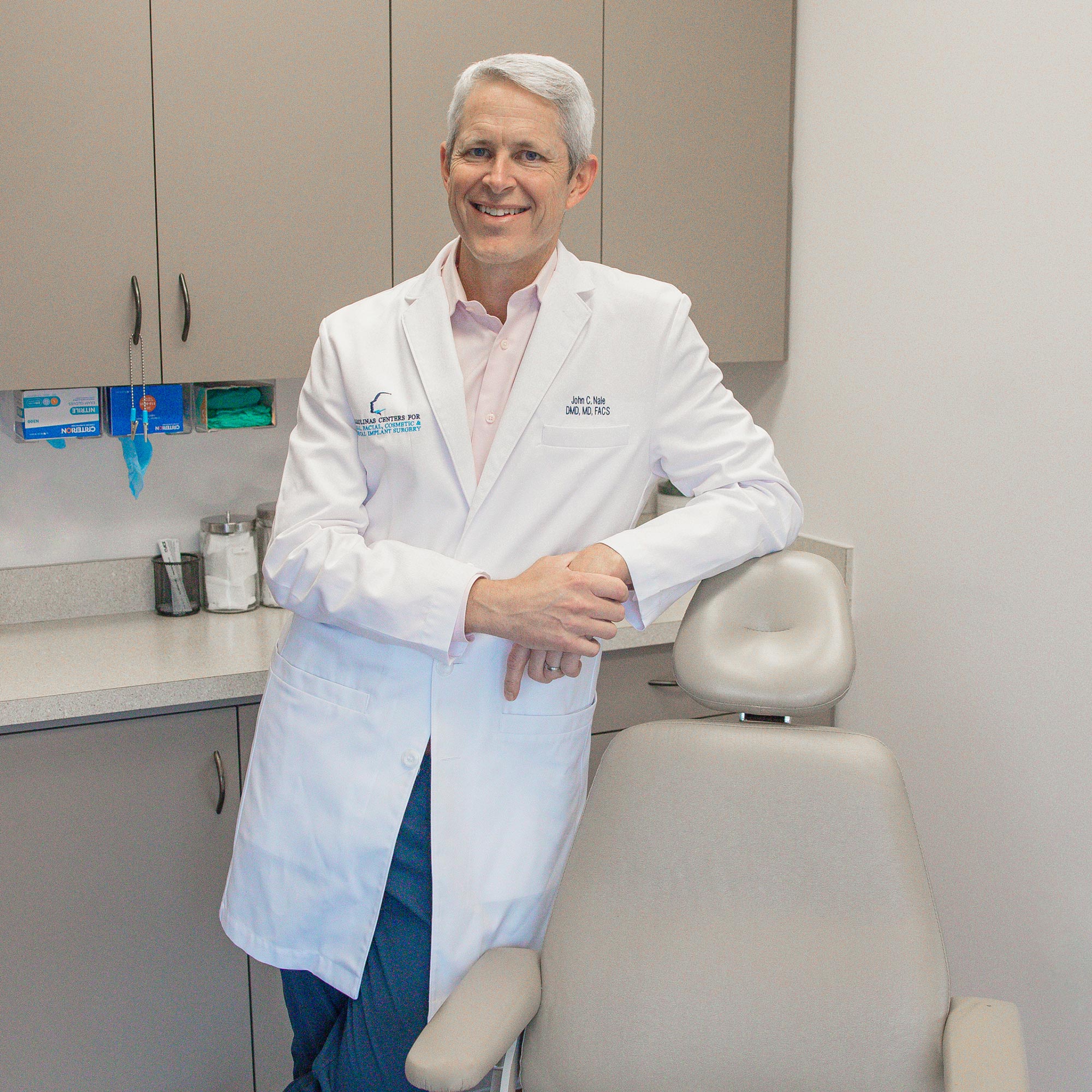 A person in a white coat stands beside a medical chair in an examination room, surrounded by cabinets and medical supplies.