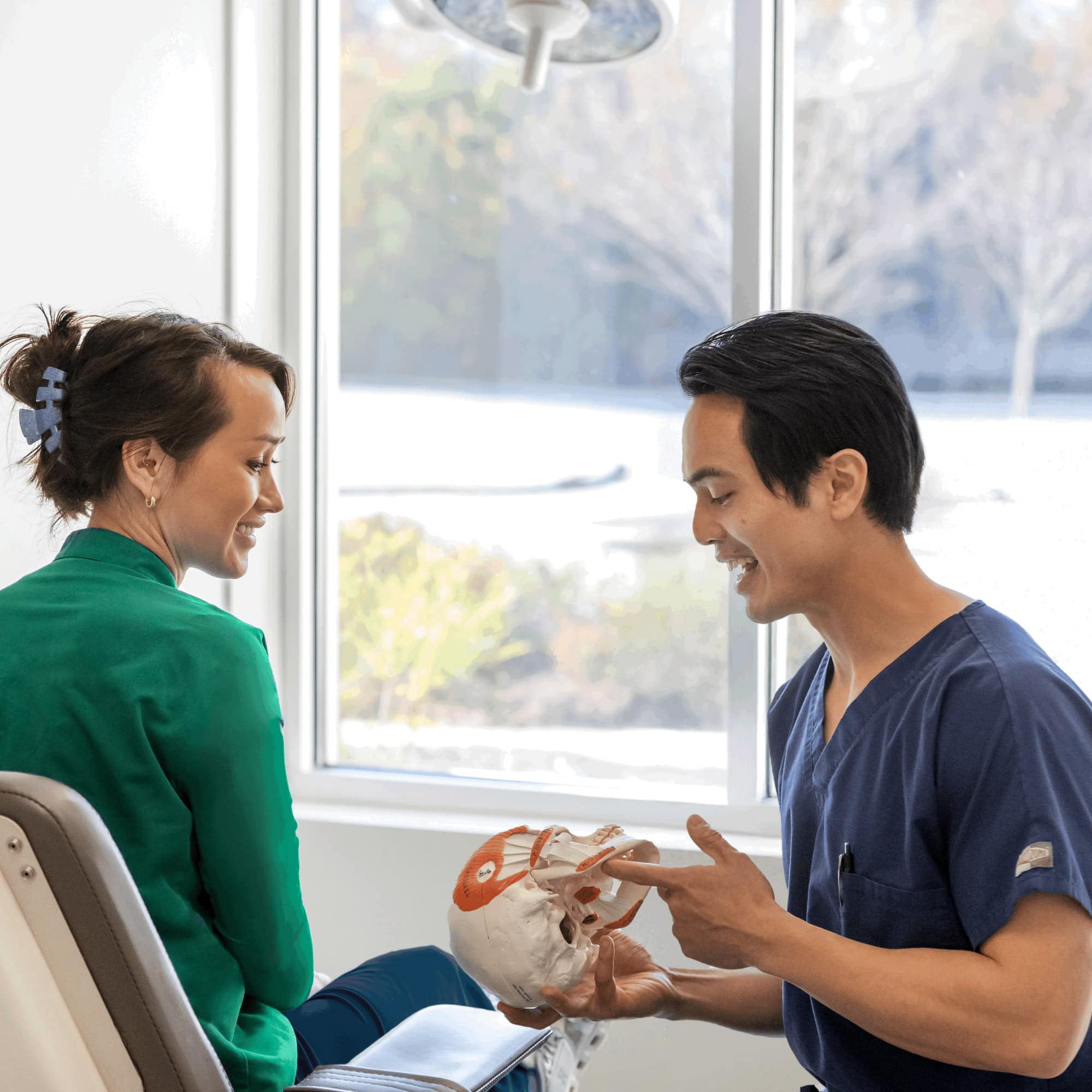 Two people in a bright room, one in scrubs holding a skull model, discussing medical or dental topics.