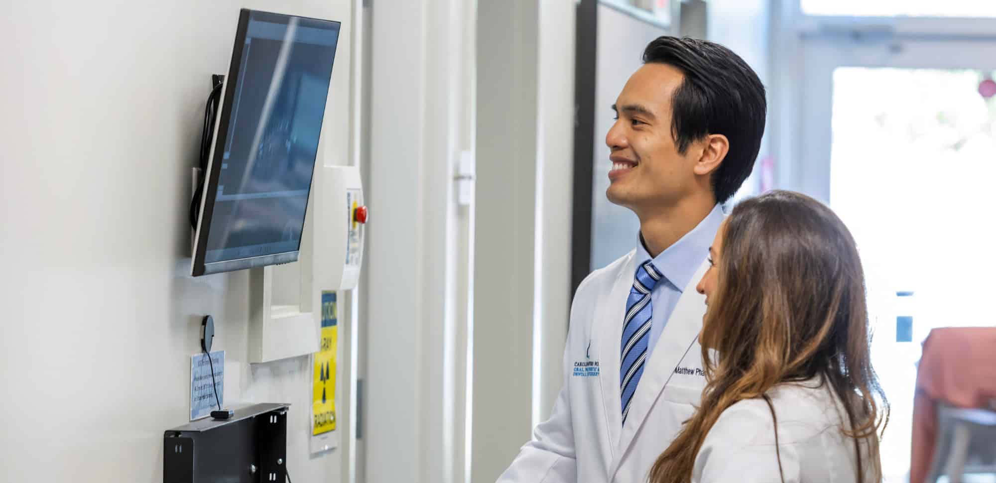 Two persons in white coats observe a mounted screen displaying an X-ray. The setting suggests a medical or dental environment.
