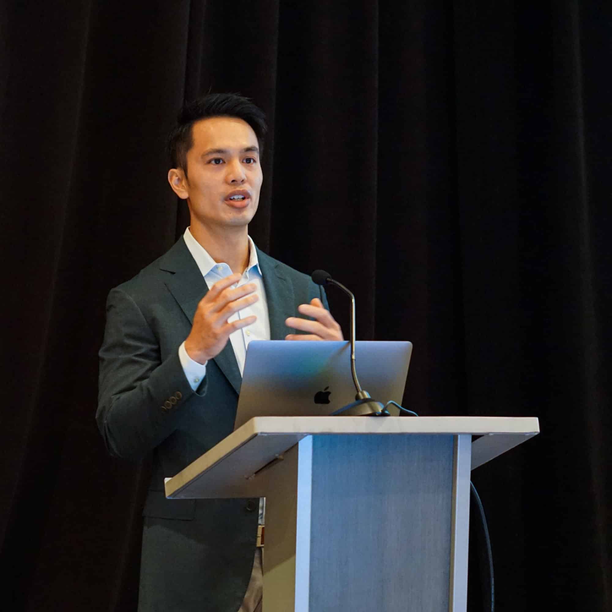 A person in a suit speaks at a podium with a laptop, against a dark curtain backdrop, during a presentation or conference.