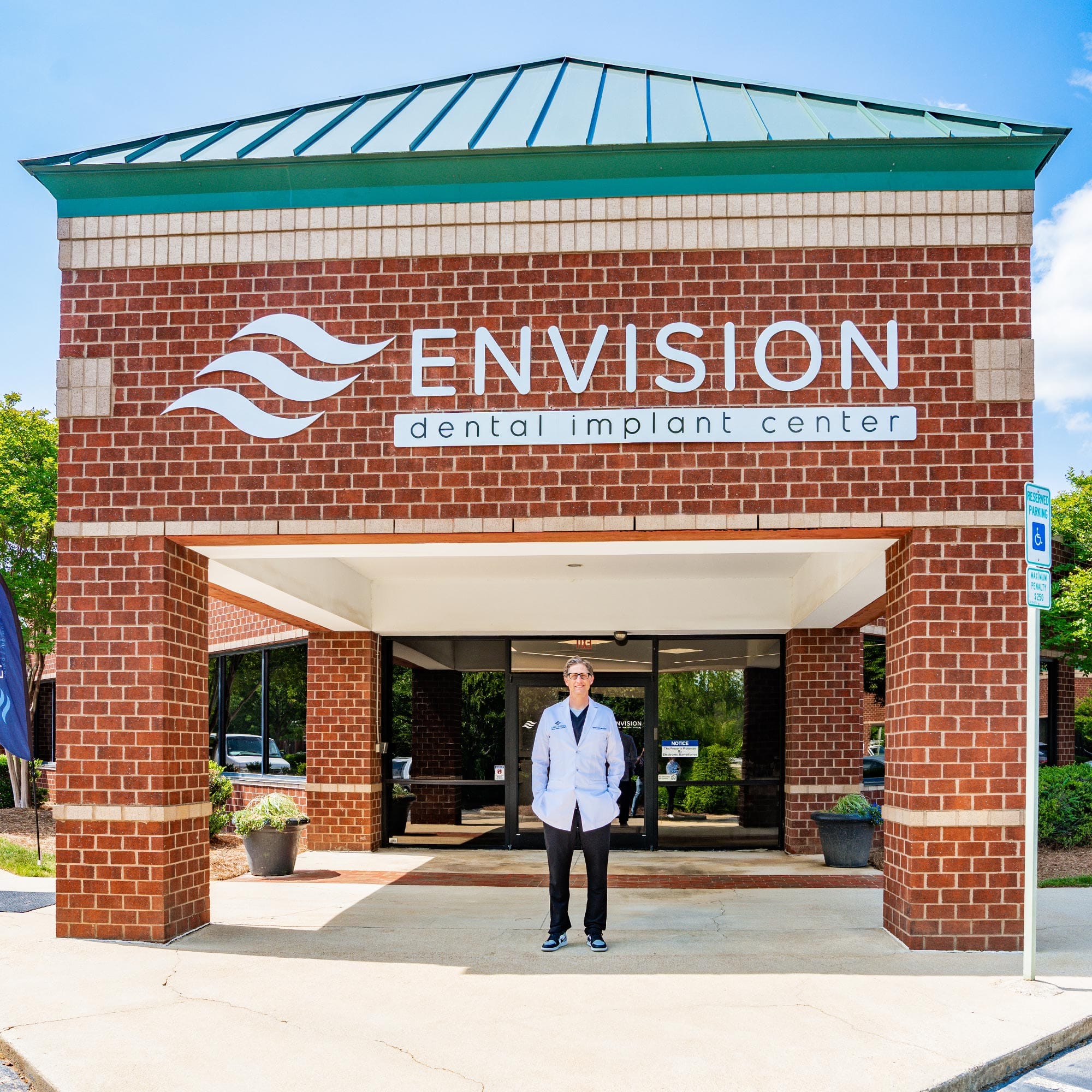 A person stands outside Envision Dental Implant Center, a brick building with green roof accents, under a sunny blue sky.