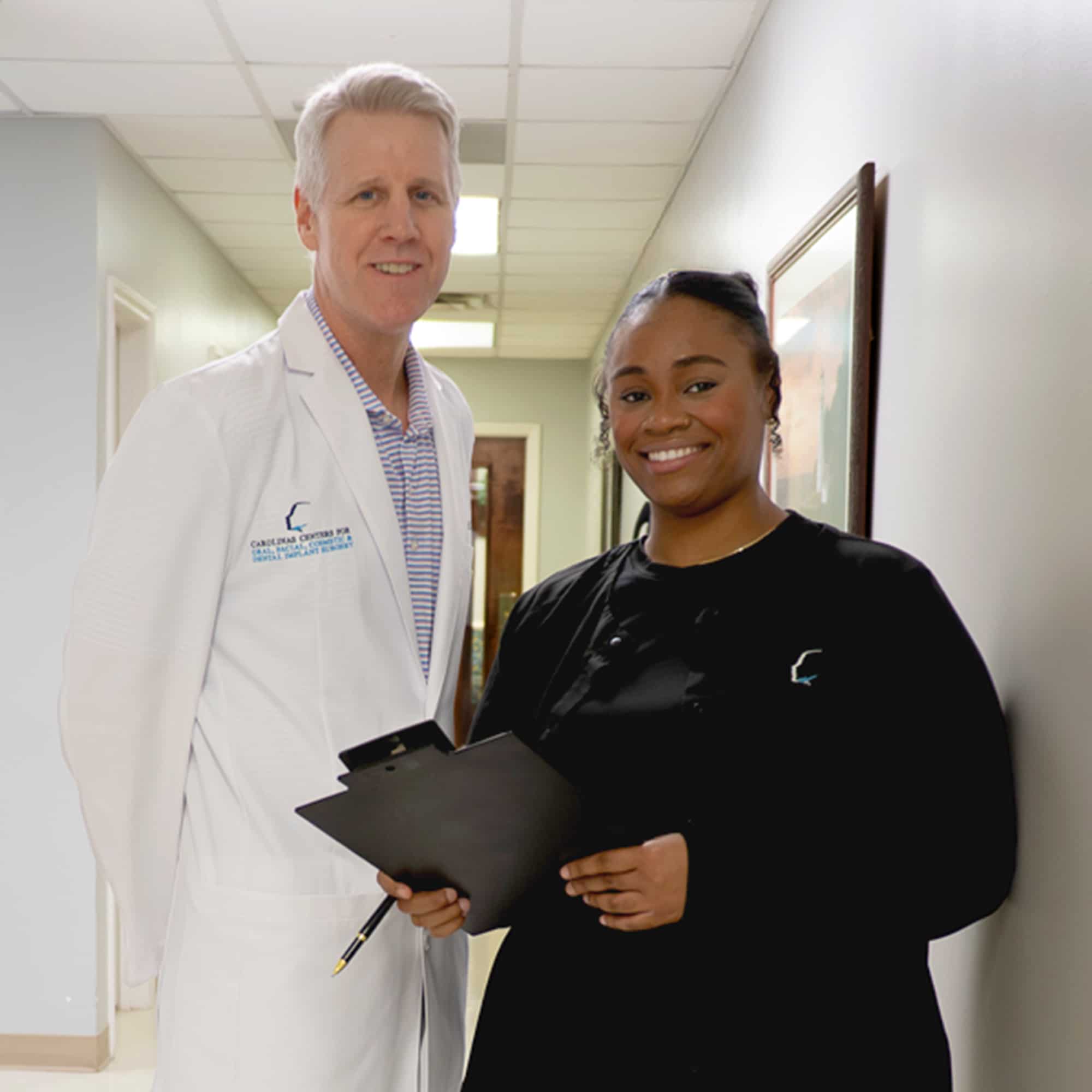 Two persons, one in a white coat and another in black attire, smiling in a hallway. Both holding clipboards.