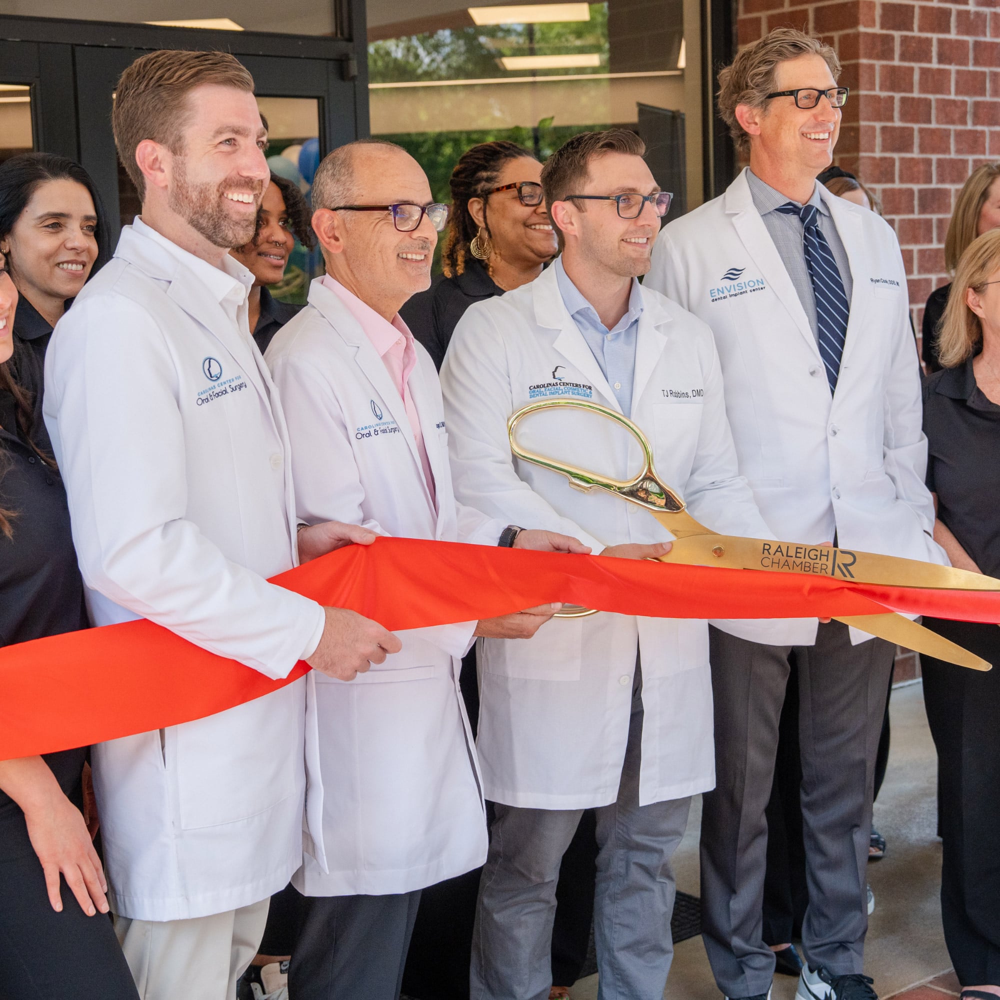 People wearing white coats participate in a ribbon-cutting ceremony, holding large scissors. The ribbon is red, and they are smiling.