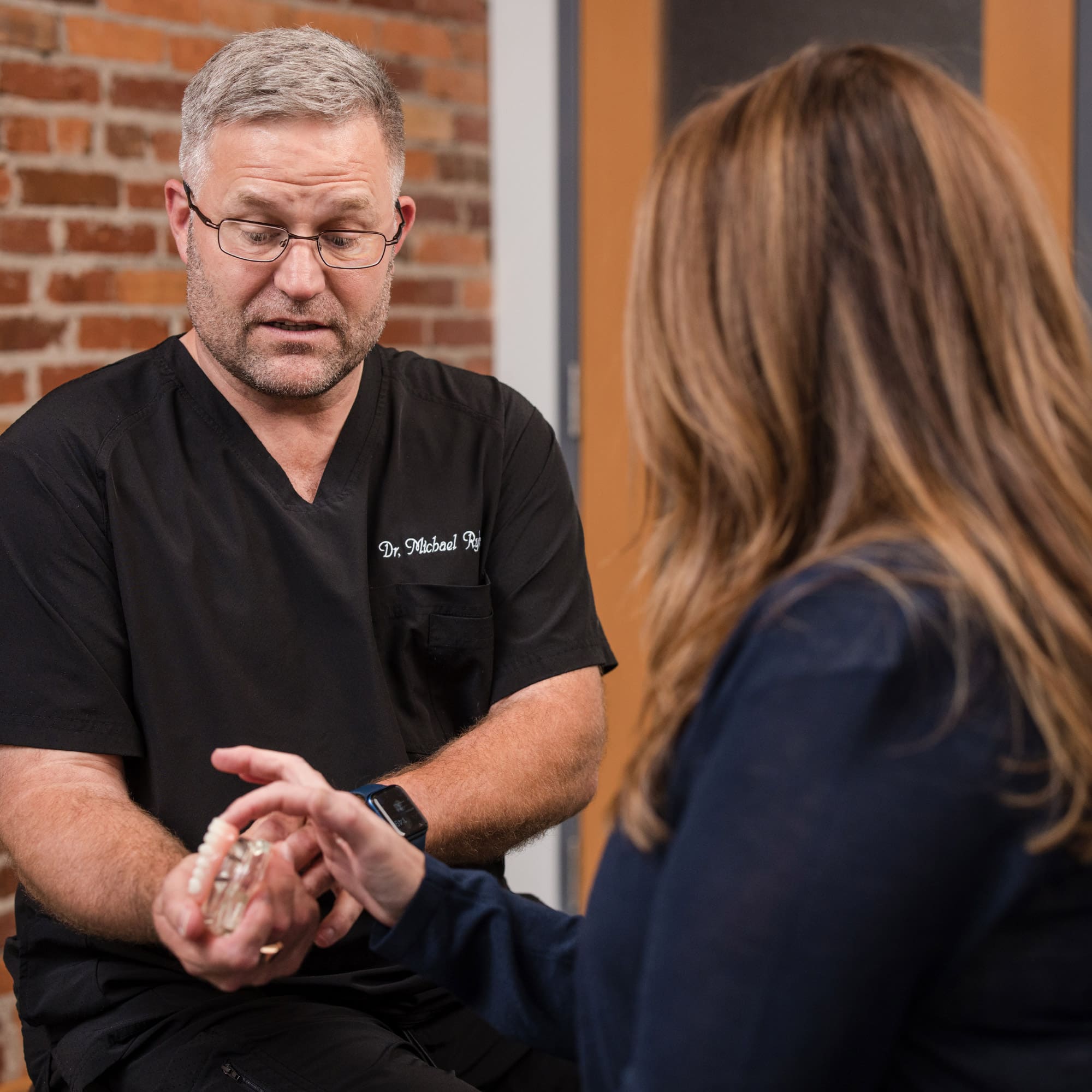A person wearing scrubs discusses a dental model with another person, set against a brick wall backdrop in an office setting.