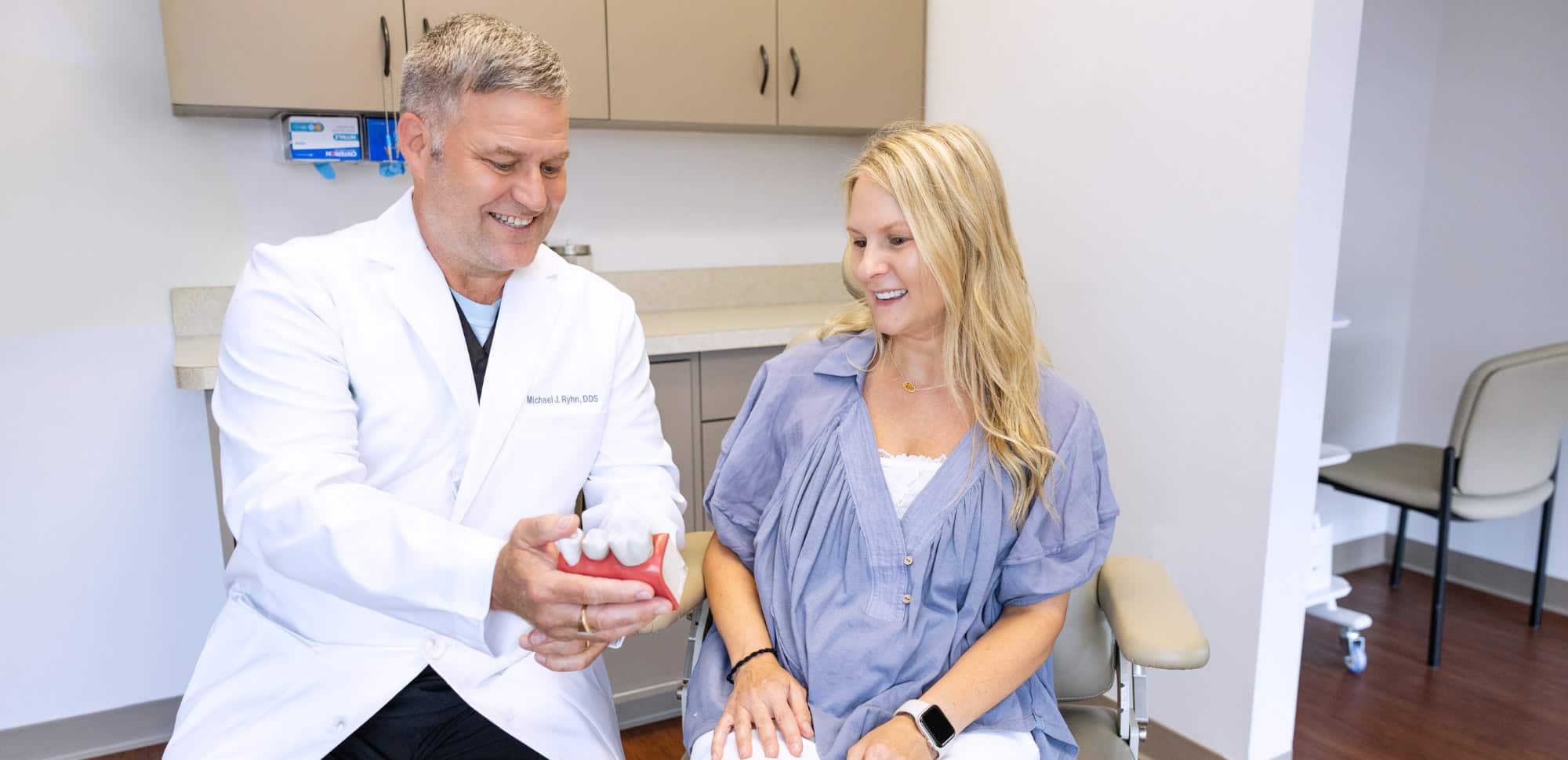 Two people are in a dental office. One person in a lab coat displays a dental model, while the other smiles and listens.