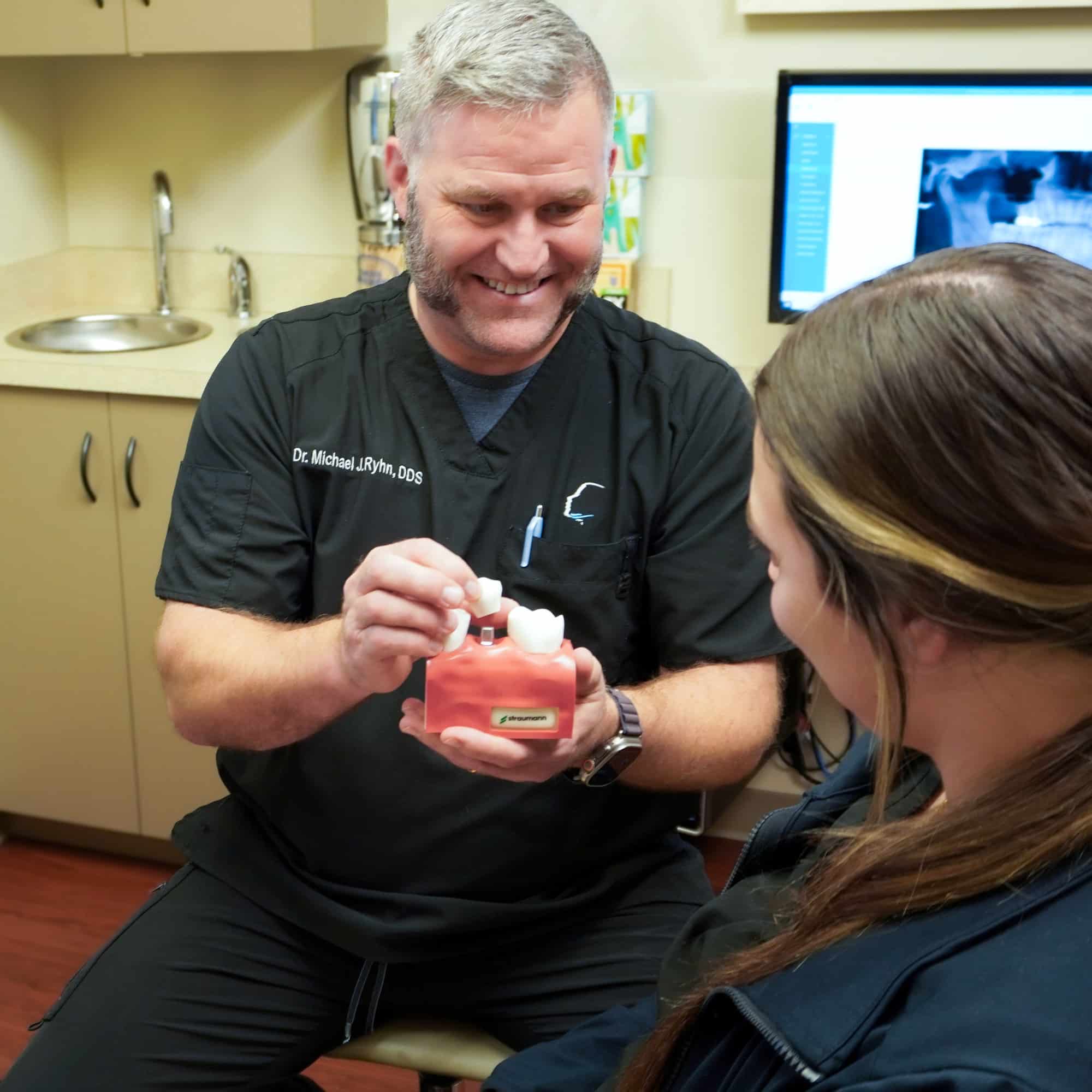 A dental professional explains a procedure to a person using a dental model in a clinic setting, with equipment visible in the background.