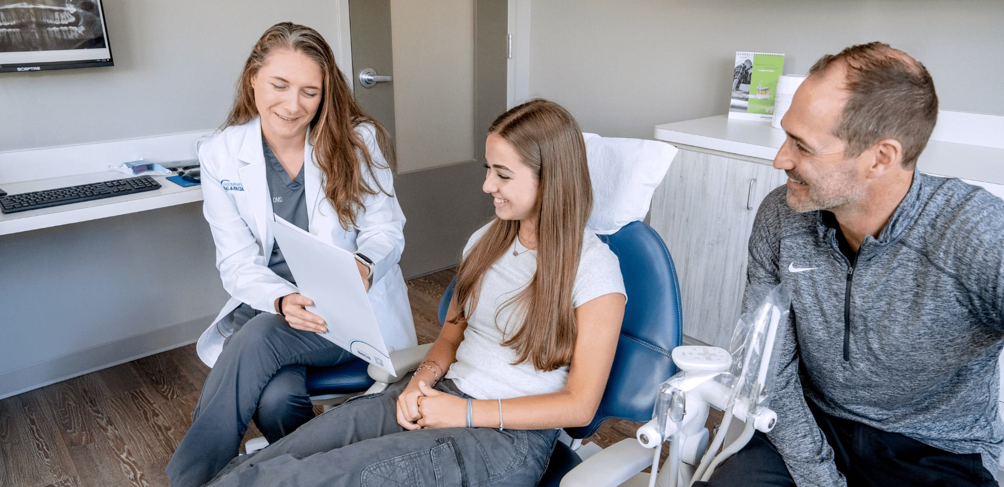 A person in a white coat discusses dental care with a young person and another adult in a modern dental office setting.