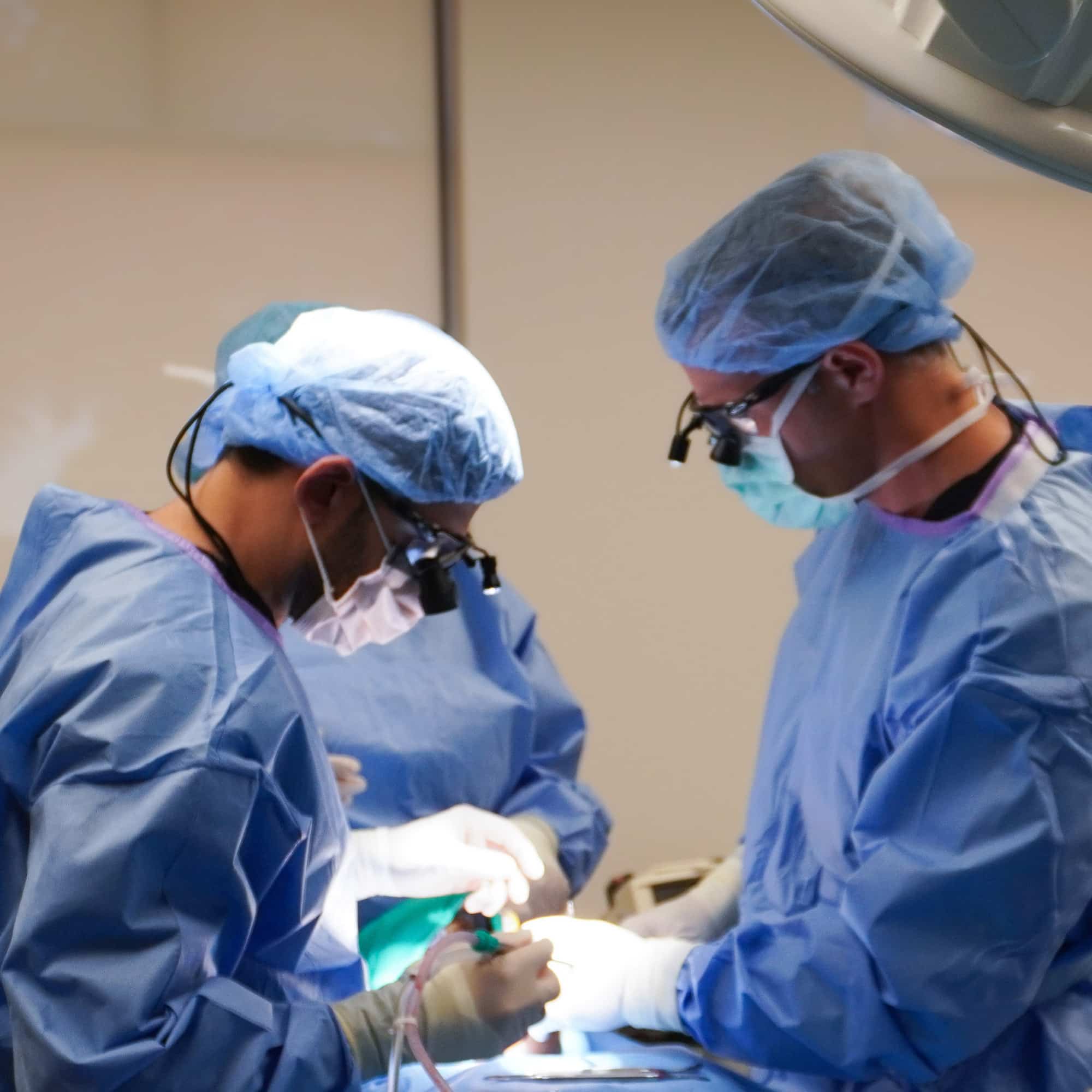 Two people in surgical gear performing a procedure under bright lights in an operating room, focused and using precision instruments.