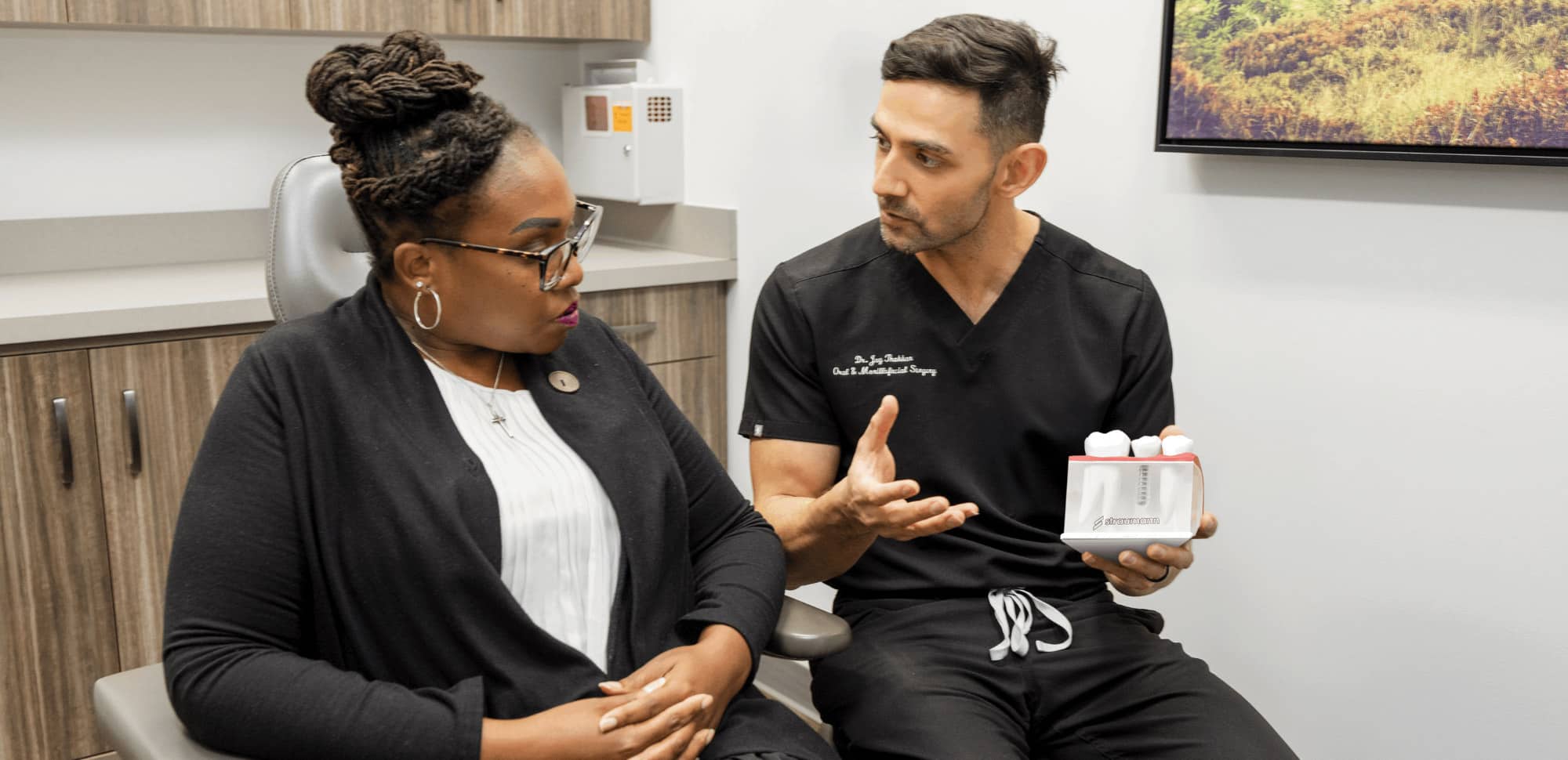 Two people in a dental office, one holding a dental model, engaged in conversation. Modern interior with wooden cabinets and framed artwork.
