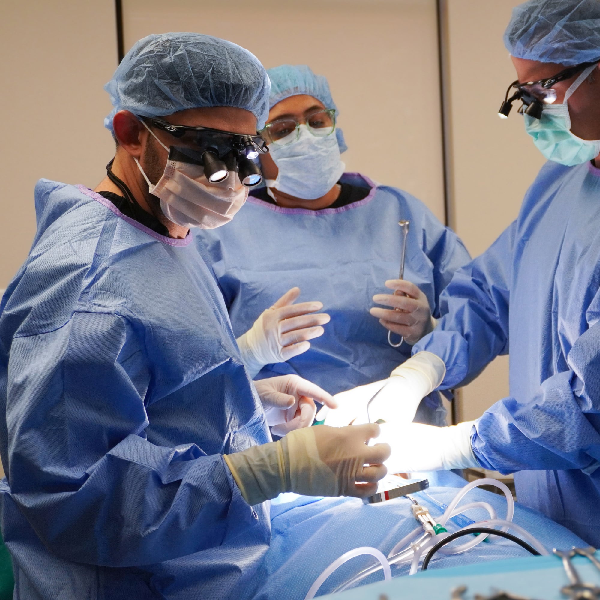 Three medical professionals in blue scrubs and masks perform surgery under bright lights in an operating room, focusing attentively on their task.
