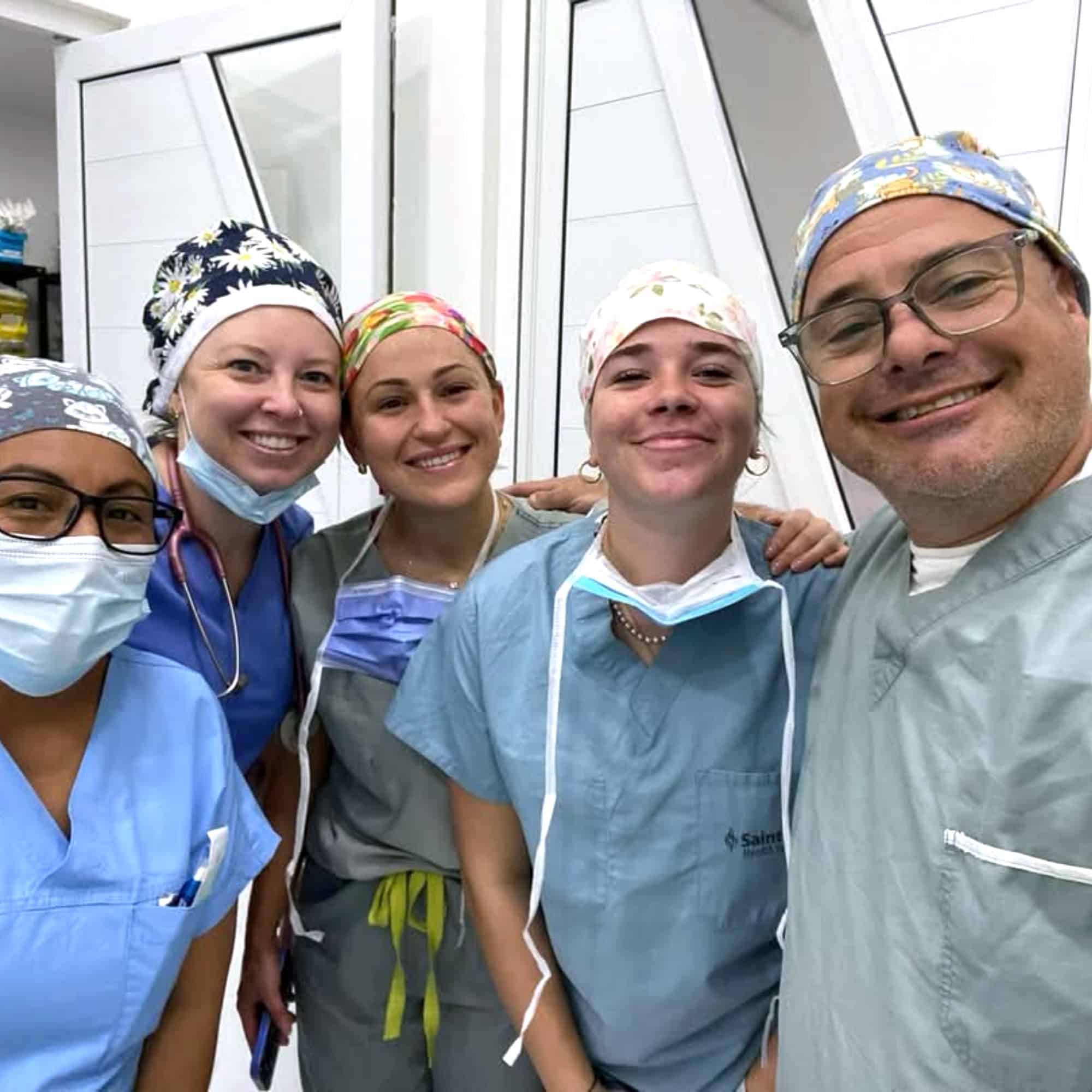Five people in scrubs, smiling and wearing surgical caps, gathered indoors against a white background. They appear to be medical professionals.