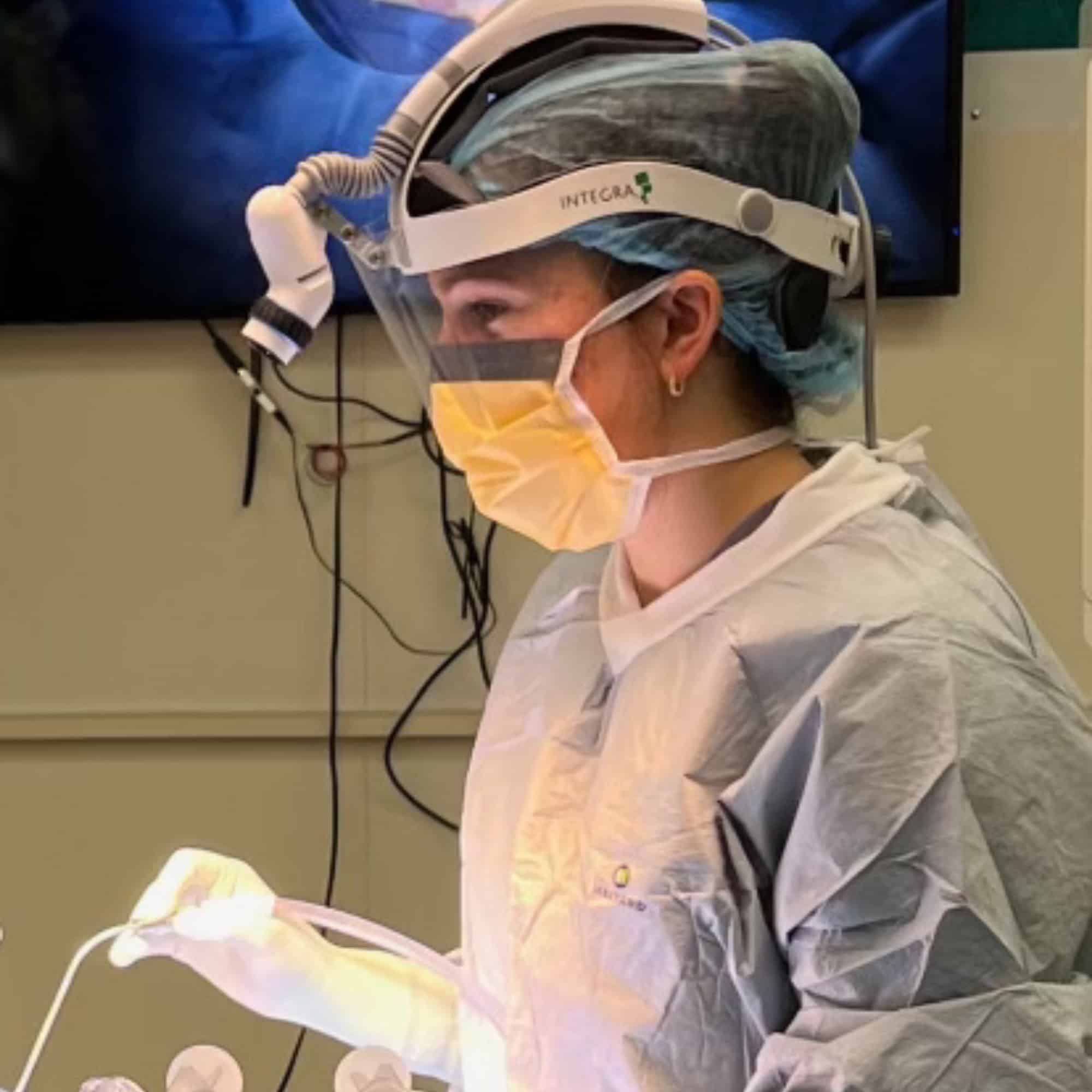 A person in surgical attire, including mask and shield, operates in a well-lit healthcare environment, focusing intently on the task.
