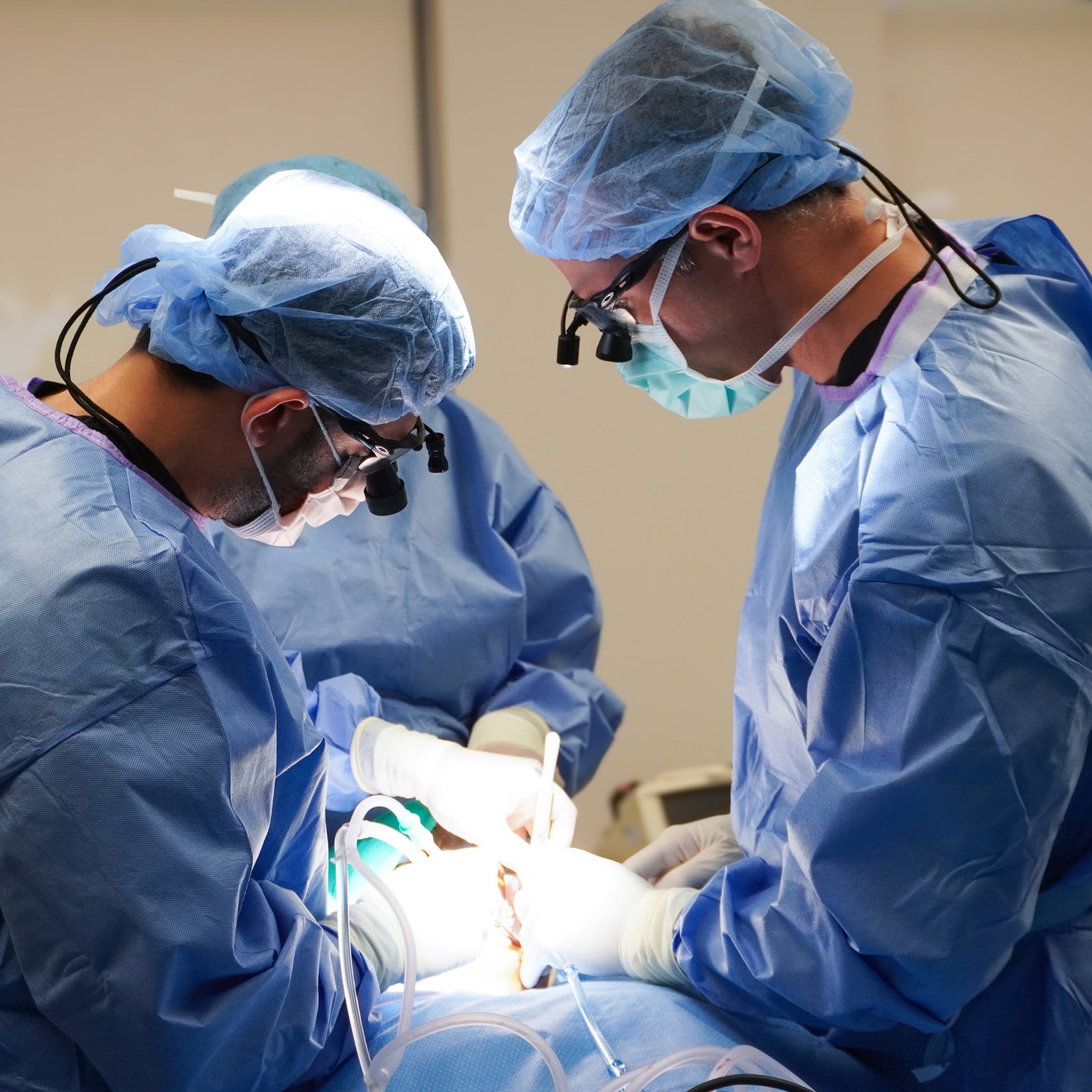 People in blue surgical attire performing an operation, focused under bright lights, with precision tools and teamwork in a medical setting.