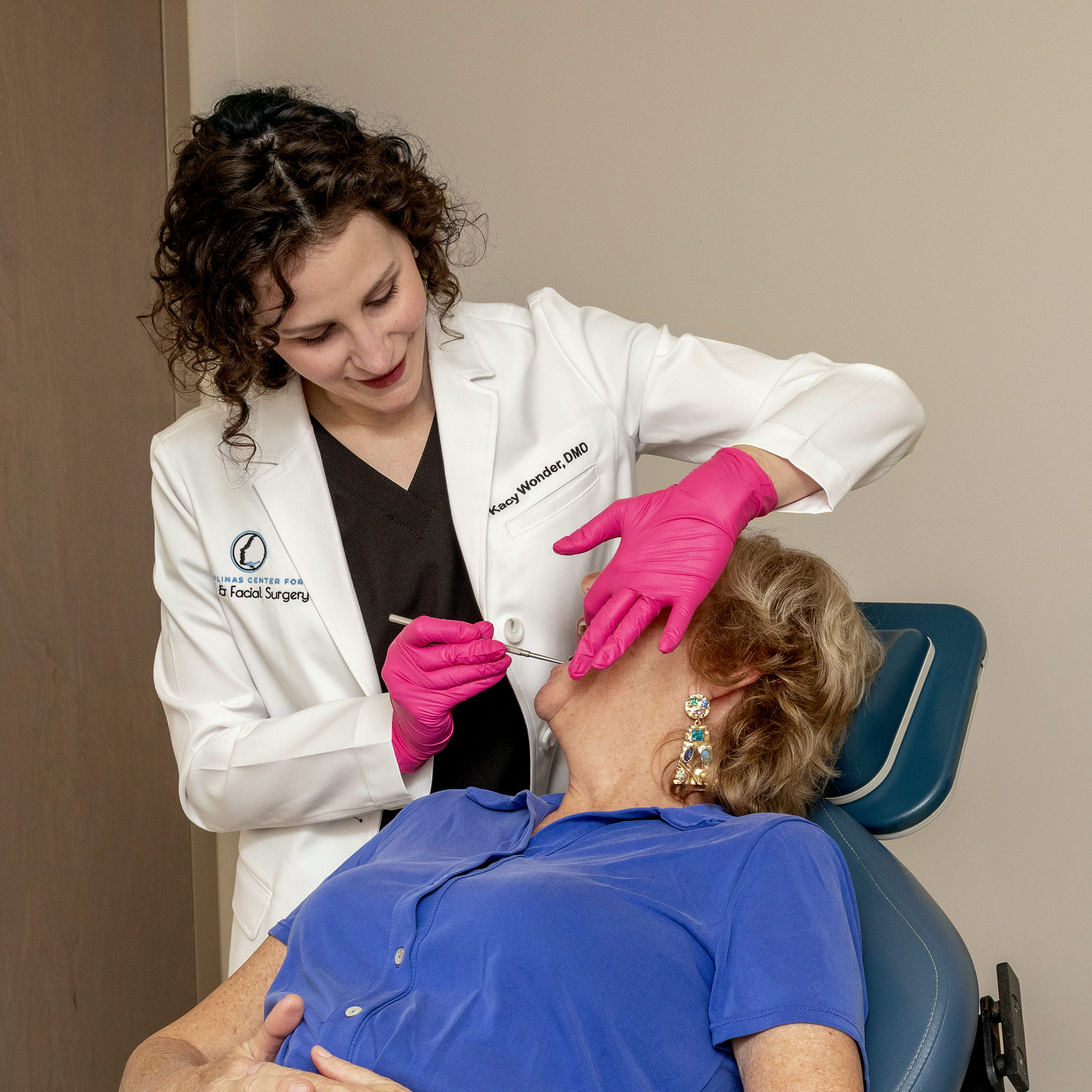 A person in a white coat examines another person in a dental chair. The environment is clinical, with medical equipment nearby.