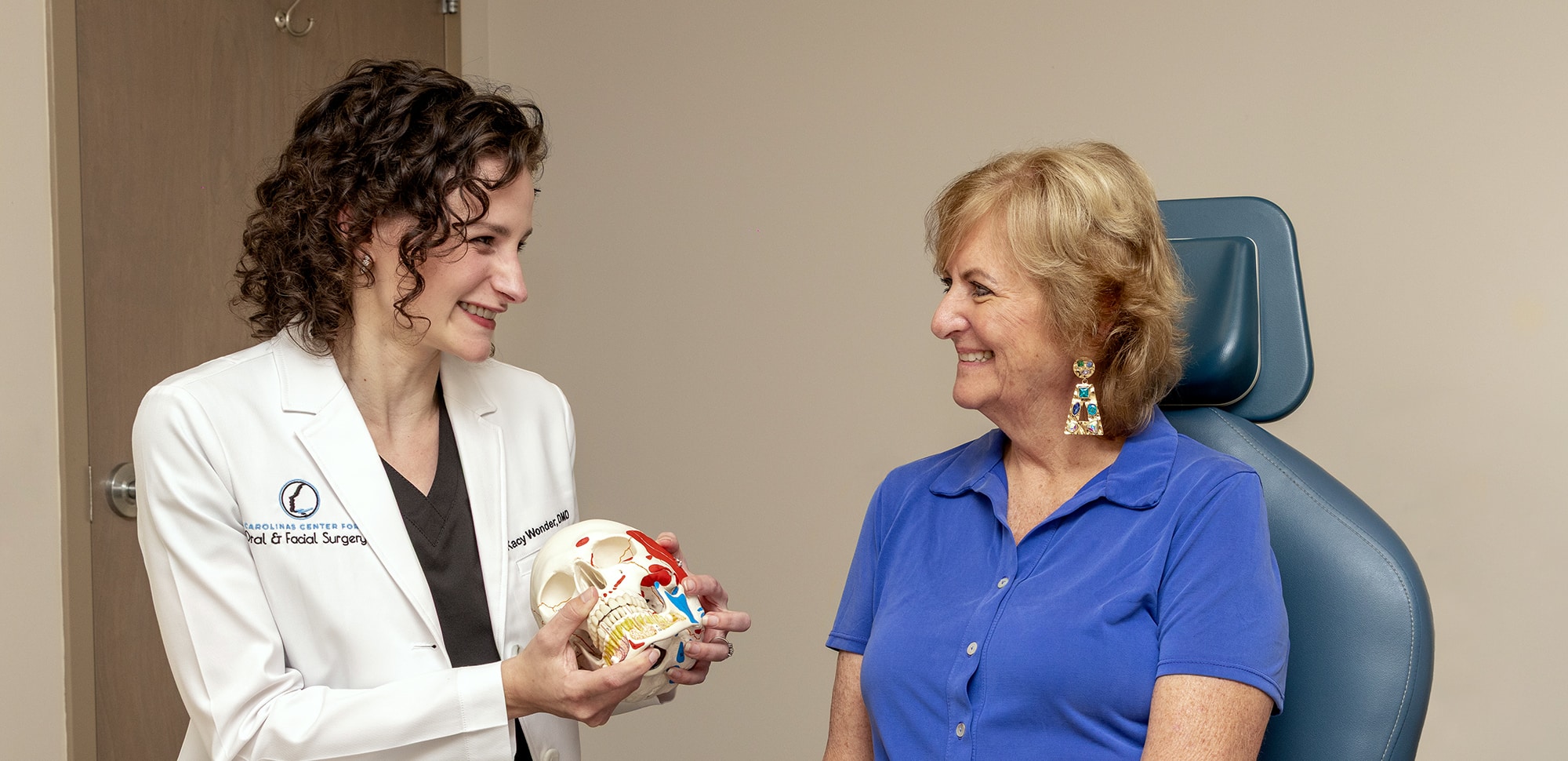 A person in a lab coat shows a skull model to another person in a medical office setting.
