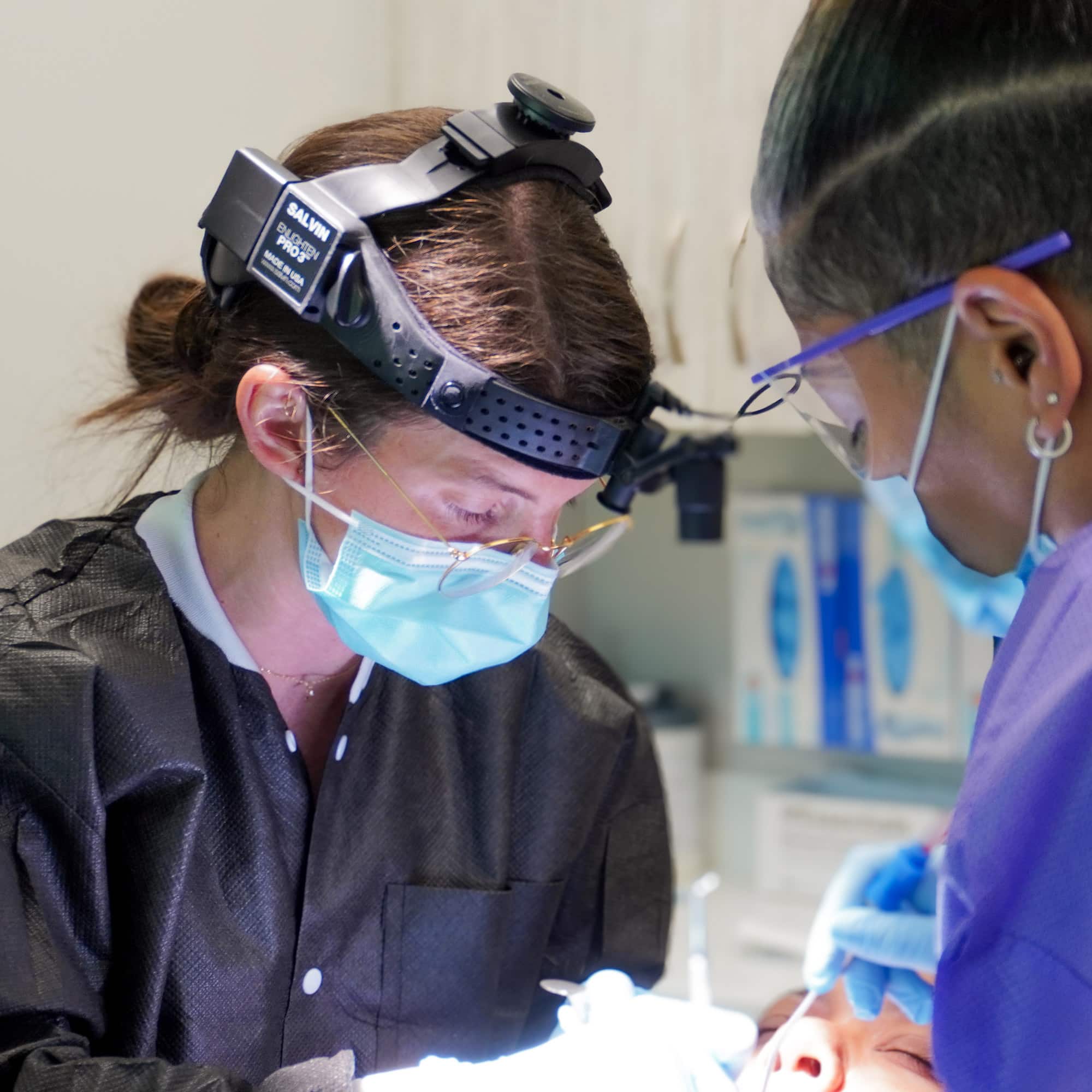 Two dental professionals, wearing masks and headlamps, perform a procedure on a patient in a brightly lit dental clinic room.