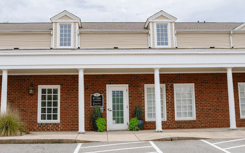 A brick building with white columns, featuring multiple windows and a central entrance. Two small shrubs flank the door, creating a tidy entrance.