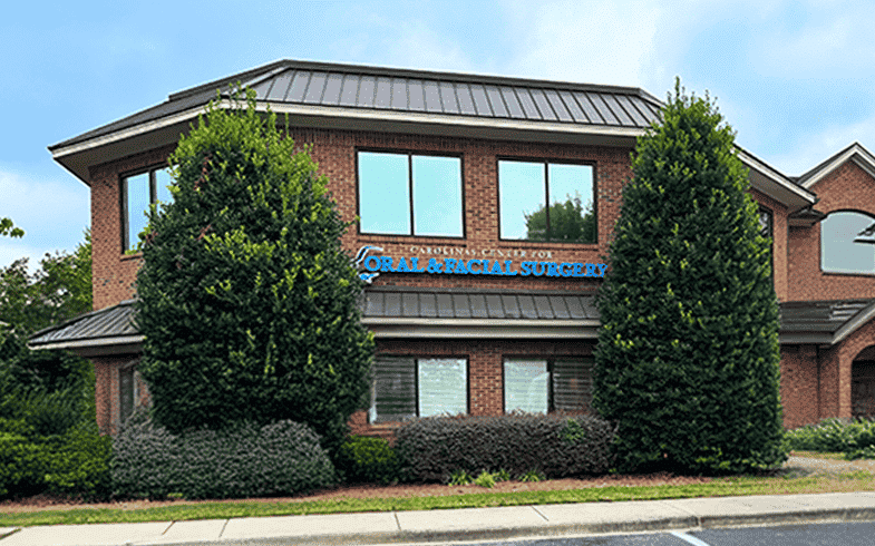 A two-story brick building with a sign for oral and facial surgery, surrounded by shrubs and trees, under a cloudy sky.