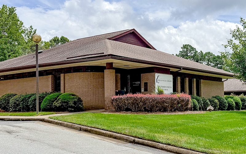 A brick building with a pitched roof, surrounded by greenery and trees, sits under a cloudy sky with a visible nameplate sign.