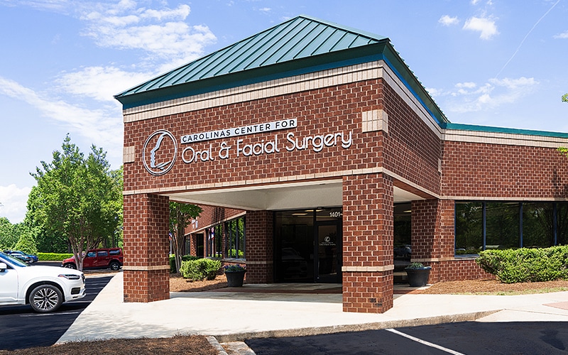 A brick building with a green roof, labeled "Carolinas Center for Oral & Facial Surgery," surrounded by parked cars and greenery under a bright sky.