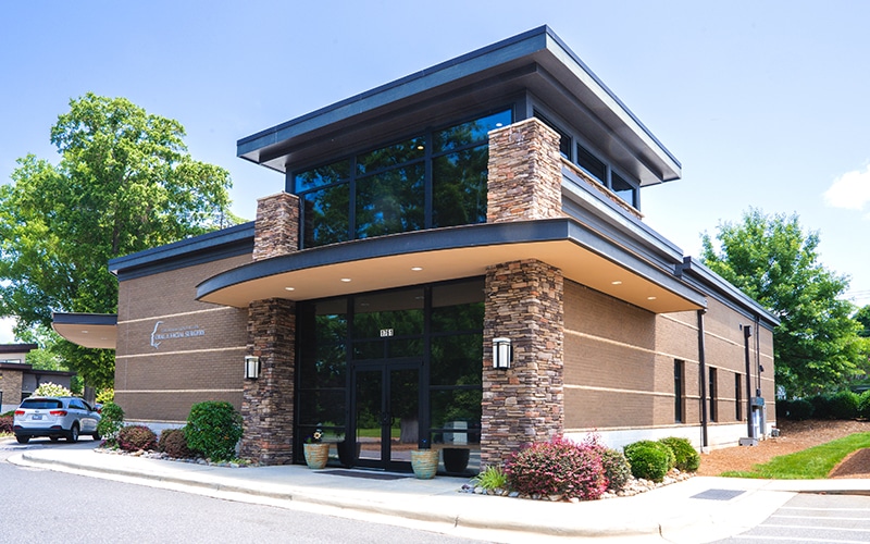 Modern single-story building with stone and brick exterior, large glass windows, surrounded by landscaping. Two parked cars and trees are visible.