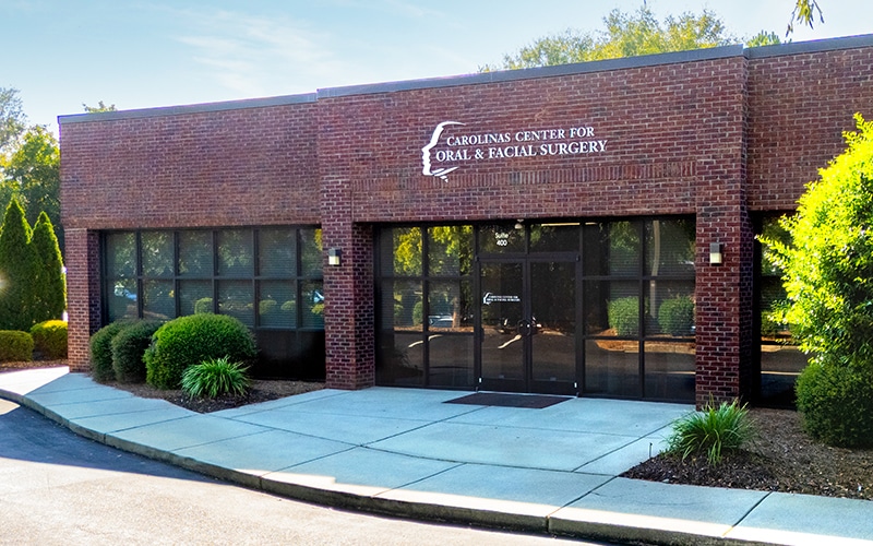A brick building with "Carolinas Center for Oral & Facial Surgery" sign, surrounded by greenery, features large windows and a glass entrance door.