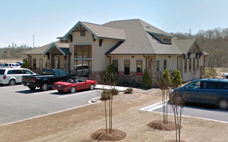 A single-story building with parked cars, trees, and a clear sky. It has a gabled roof and stone accents on the exterior walls.