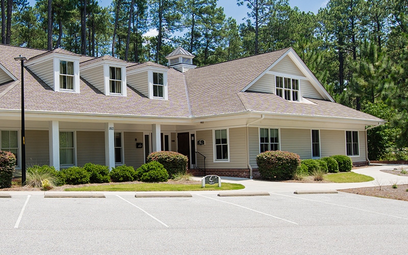 A beige house with numerous windows, surrounded by trees, sits adjacent to an empty parking lot under a clear blue sky.