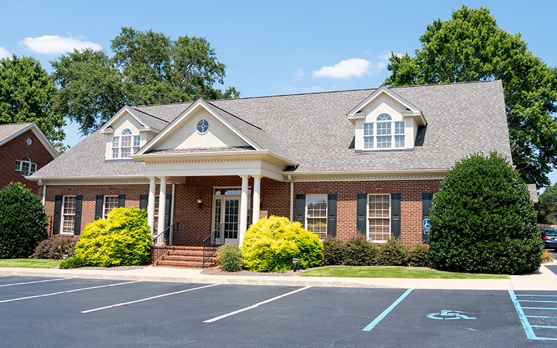 A brick building with symmetrical windows, columns, and a gabled roof is surrounded by bushes and a parking lot on a sunny day.