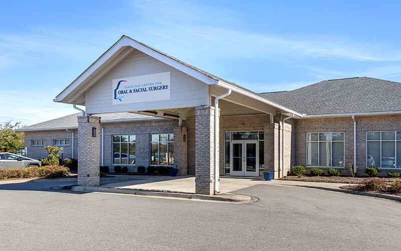 A brick building with a gable roof houses an oral and facial surgery center. The entrance is covered, with clear skies above.
