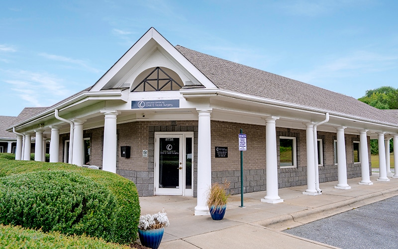 Single-story brick building with white columns, housing an oral and maxillofacial surgery office. Surrounded by trimmed bushes, under a clear blue sky.