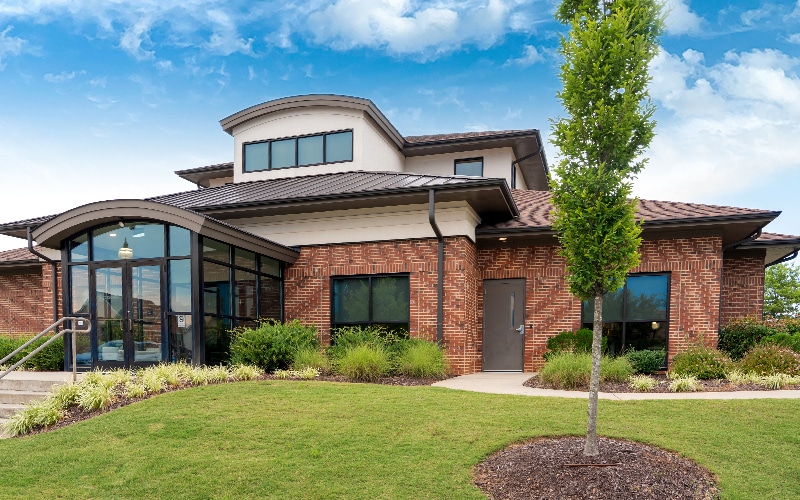 Modern brick building with large windows, surrounded by landscaped grass and plants. A clear blue sky frames the structure. No people are visible.