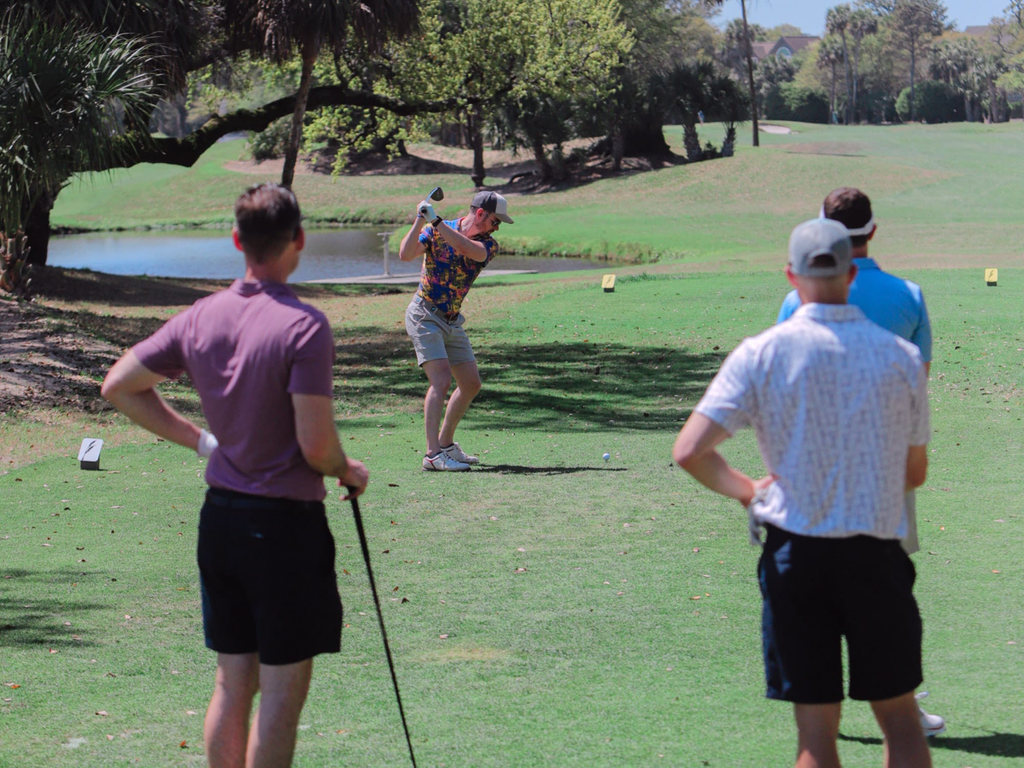 Four people on a golf course, one swinging a club. Trees and a small pond in the background, under clear skies.