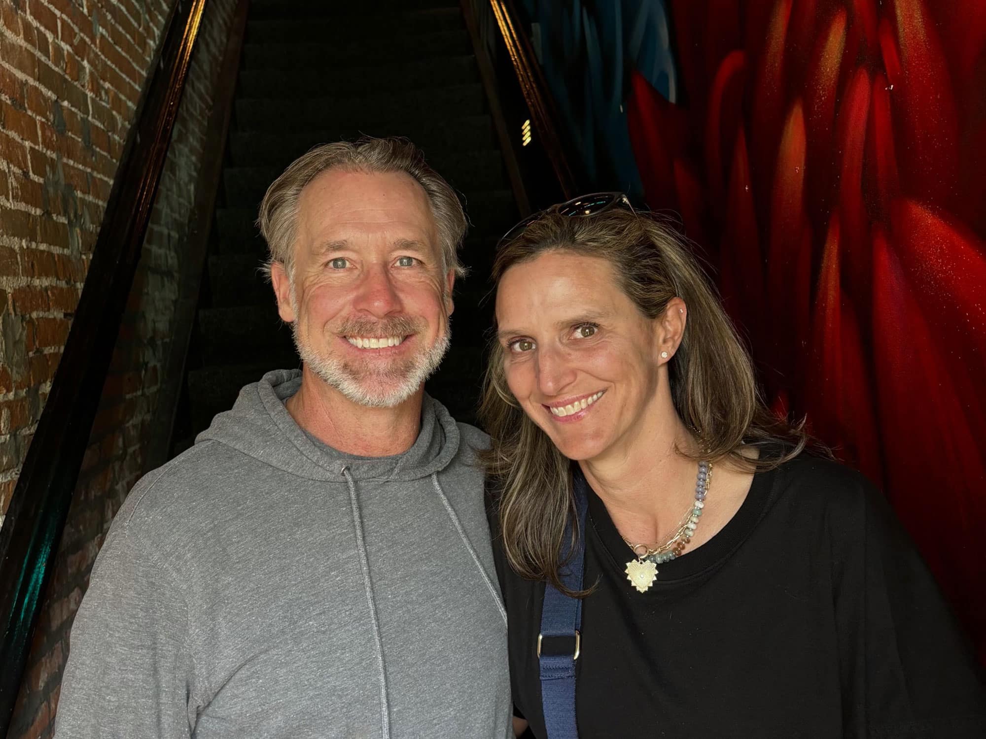 Two people smiling at the camera indoors, standing on a staircase with brick walls and vibrant artwork in the background.