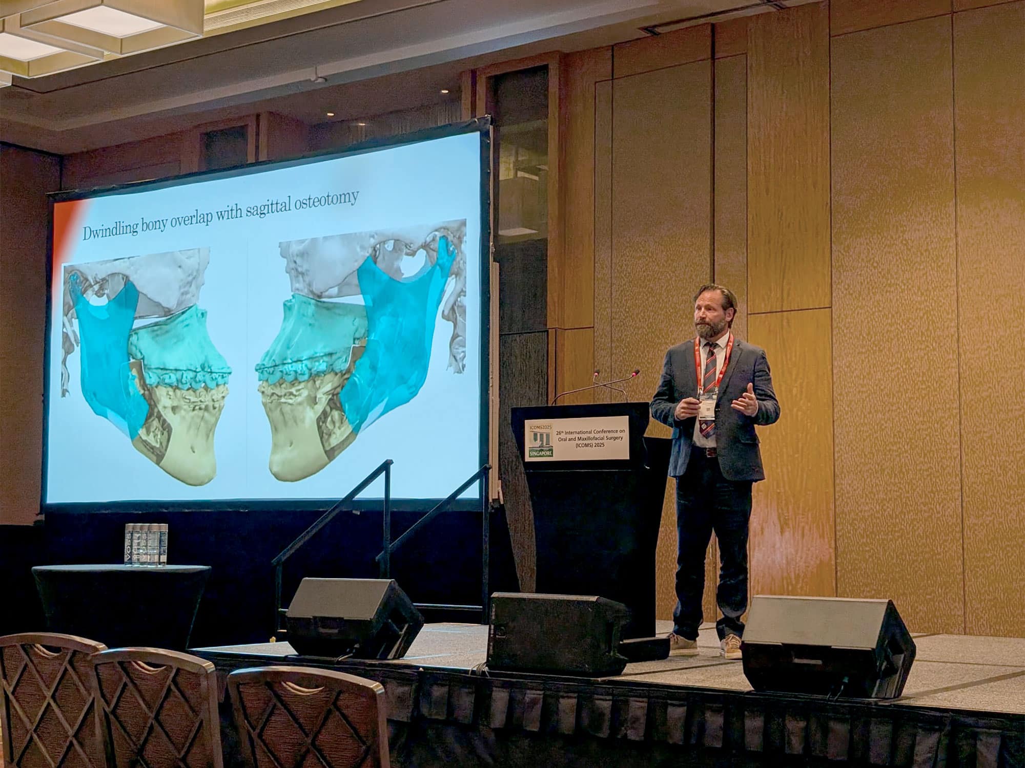 A person presents at a conference, standing beside a projected slide of bony overlap images. The podium displays conference branding.
