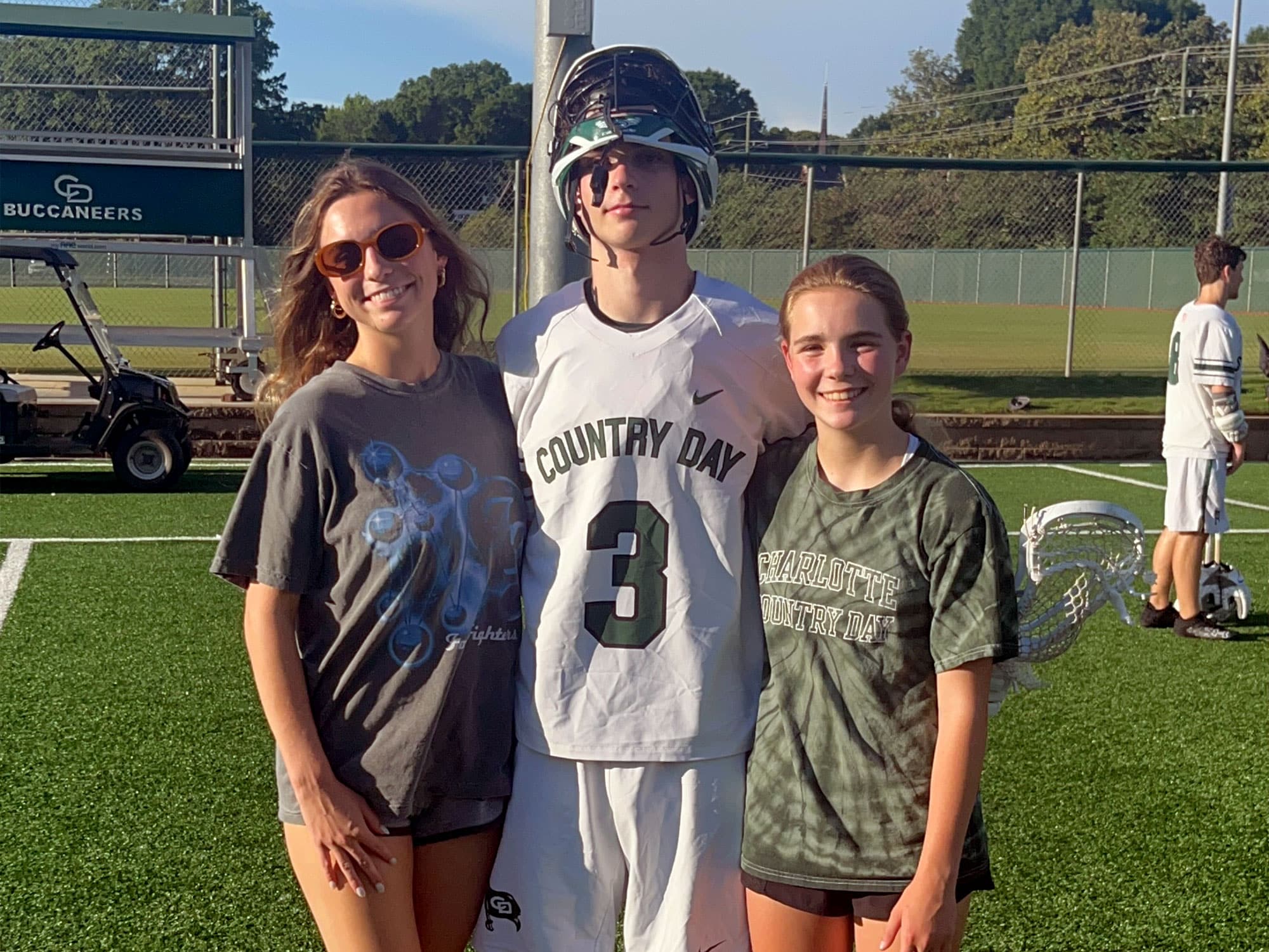 Three people standing on a sports field, one in a lacrosse uniform, with "Country Day" printed. Green fence and trees in the background.