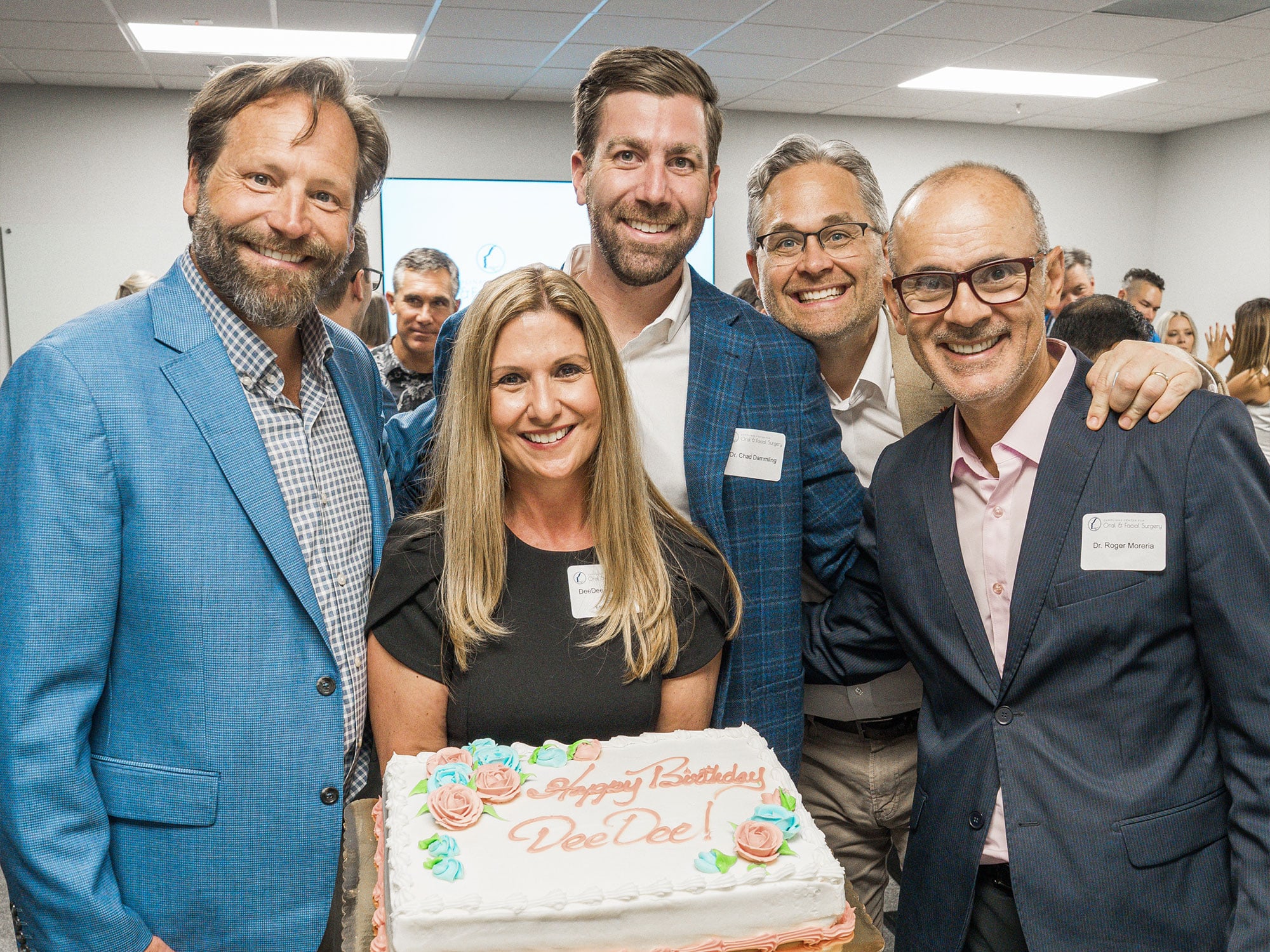 Five smiling people celebrate a birthday indoors with a frosted cake that says "Happy Birthday Dee Dee." They are gathered closely and appear joyful.